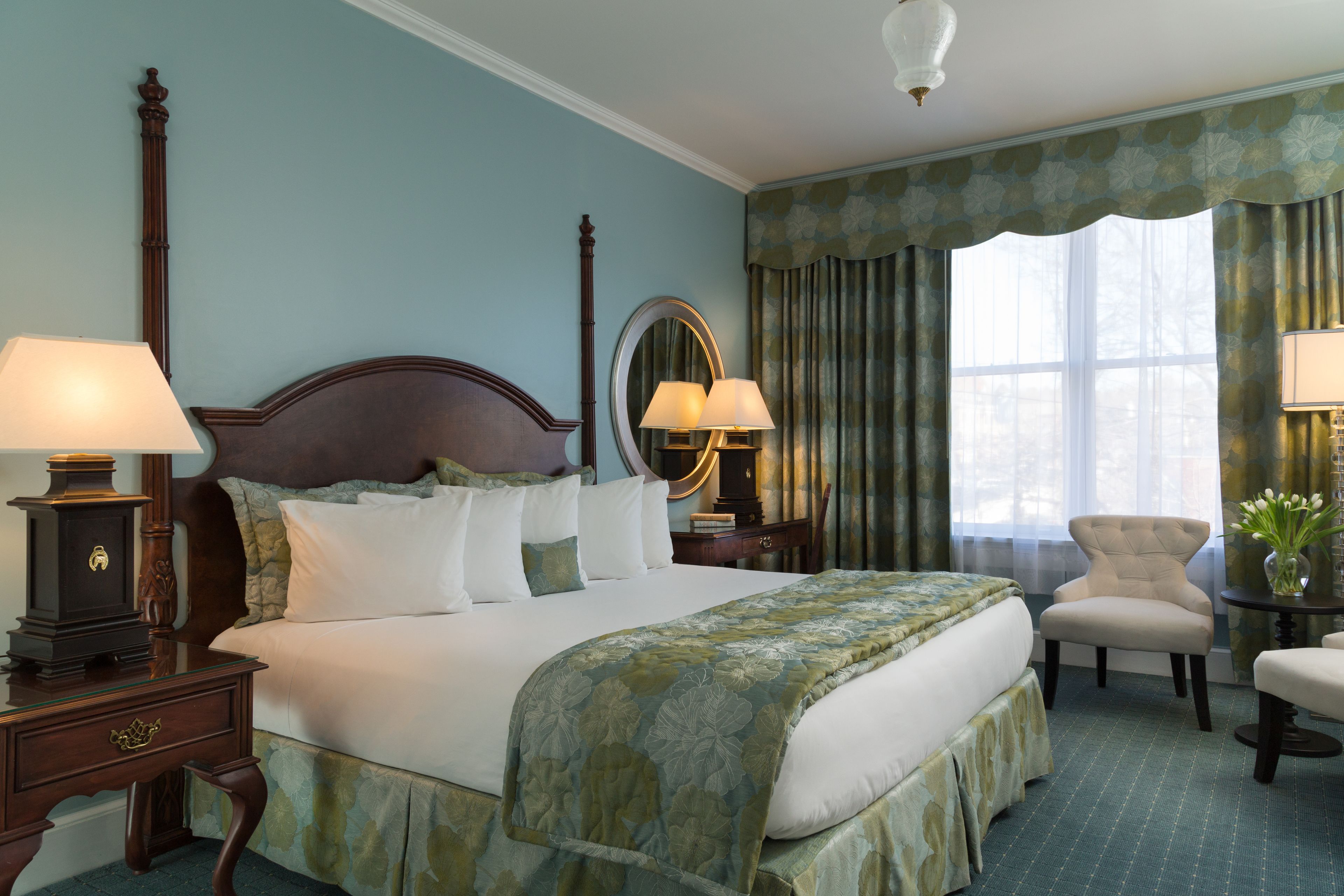 A neatly arranged hotel room with a large bed, two bedside tables with lamps, a chair, a small table with a vase, and a window with curtains.