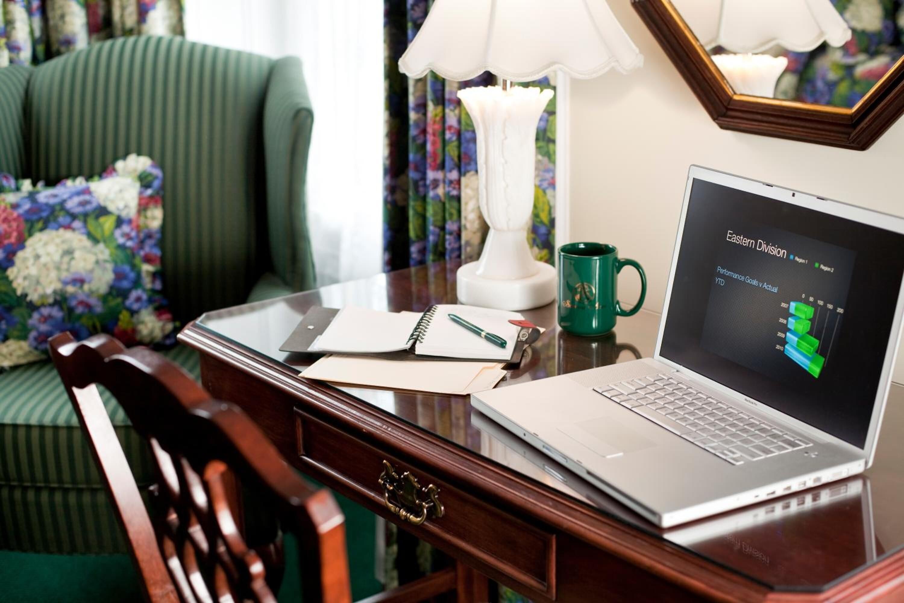 A laptop on a wooden desk with a notebook, pen, and a green mug.