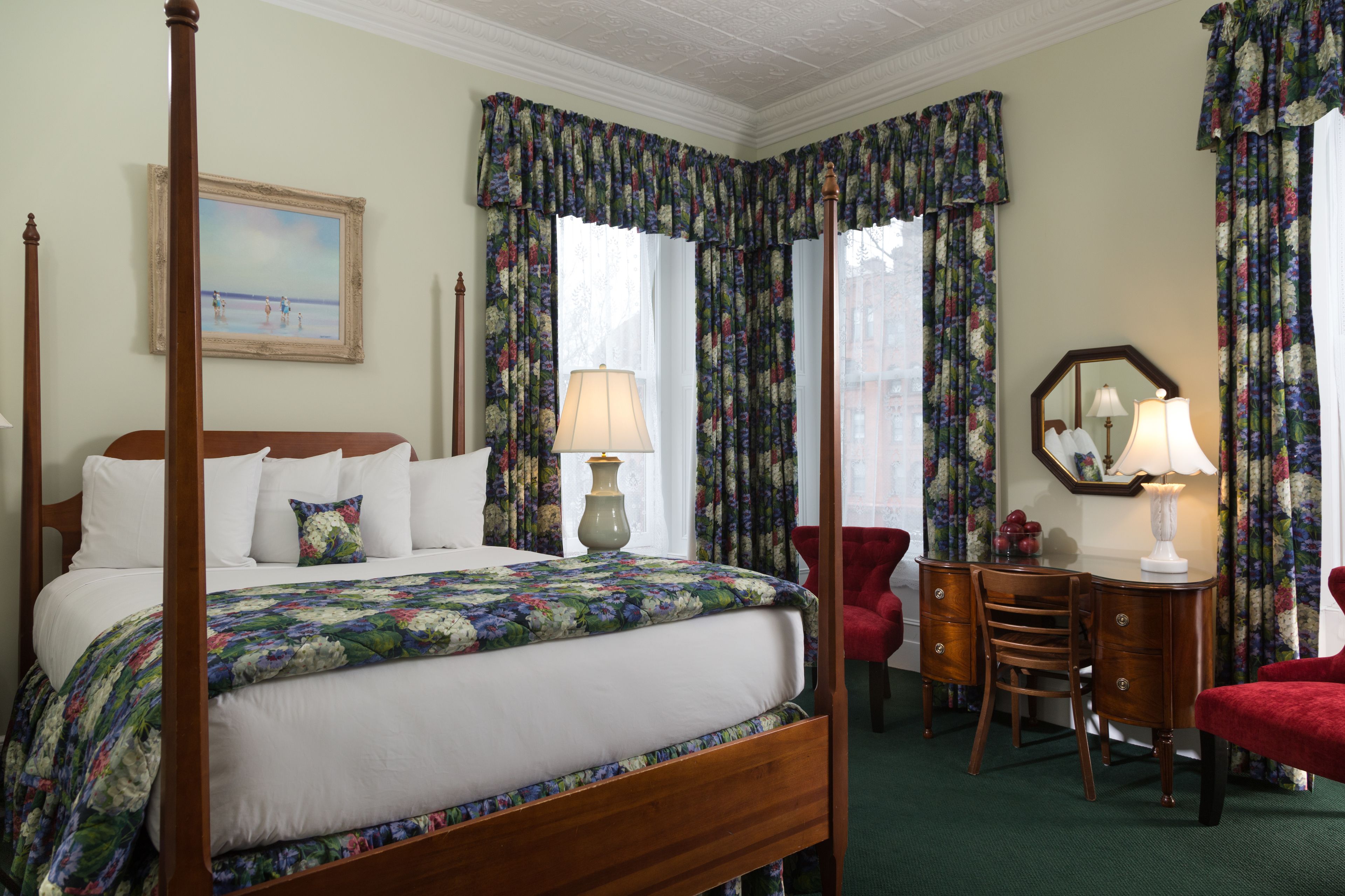 Una cama con dosel con sábanas blancas, una colcha floral y una mesita de noche con lámpara. Hay un escritorio con silla y un espejo, y un sillón rojo.