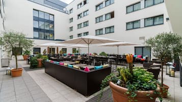 Un área de estar al aire libre con varias mesas y sombrillas, rodeada de plantas en macetas y un edificio moderno al fondo.