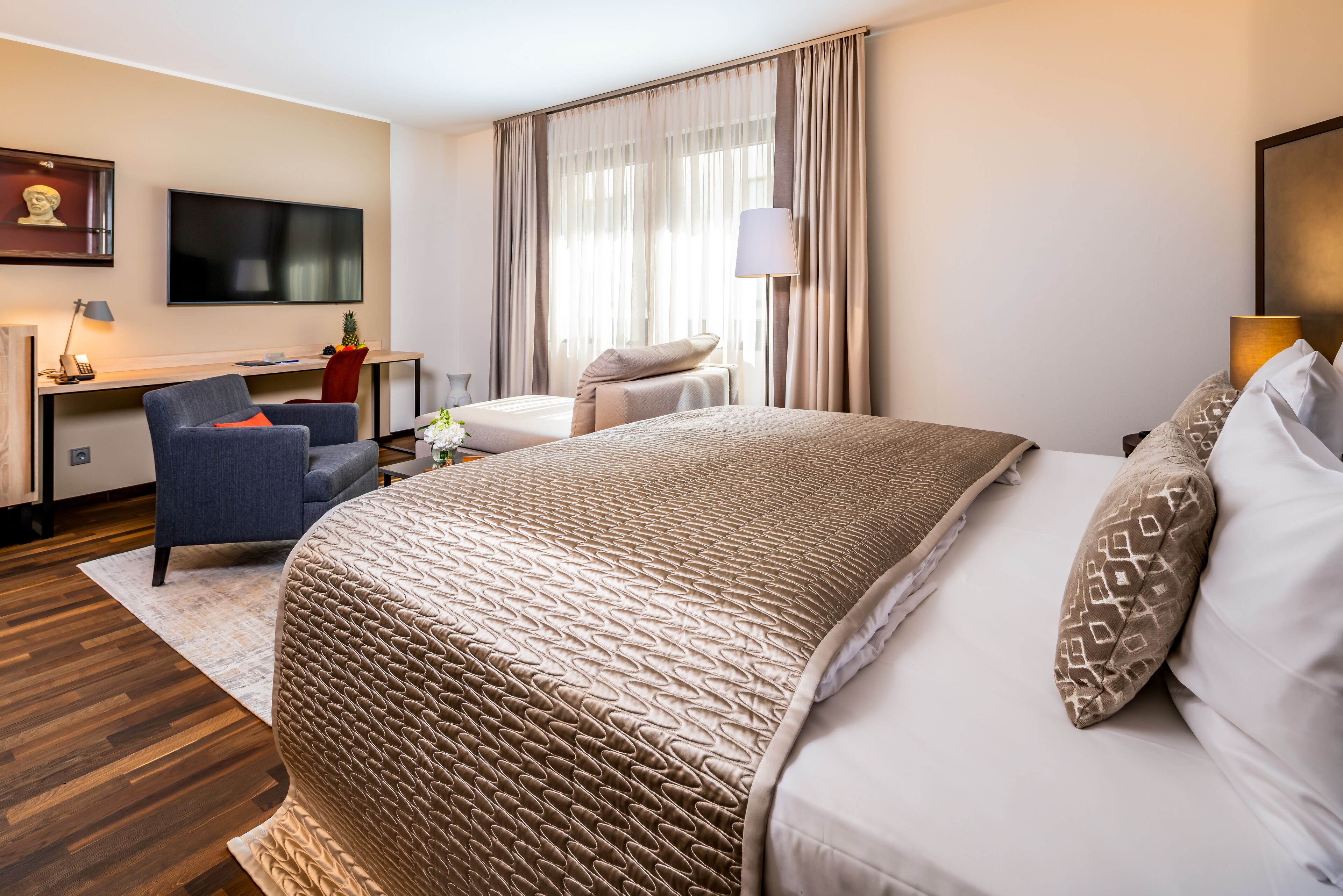 A hotel room with a large bed, a desk with a chair, a TV mounted on the wall, and a window with curtains.