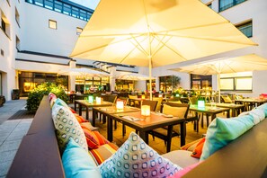 An outdoor dining area with multiple tables and chairs, large umbrellas, and cushioned seating. - Park Plaza Trier (Trier)