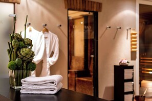 A stack of white towels on a table, a vase with green plants, and a reflection of a room with a wooden door and a hanging robe. - Park Plaza Trier (Trier)