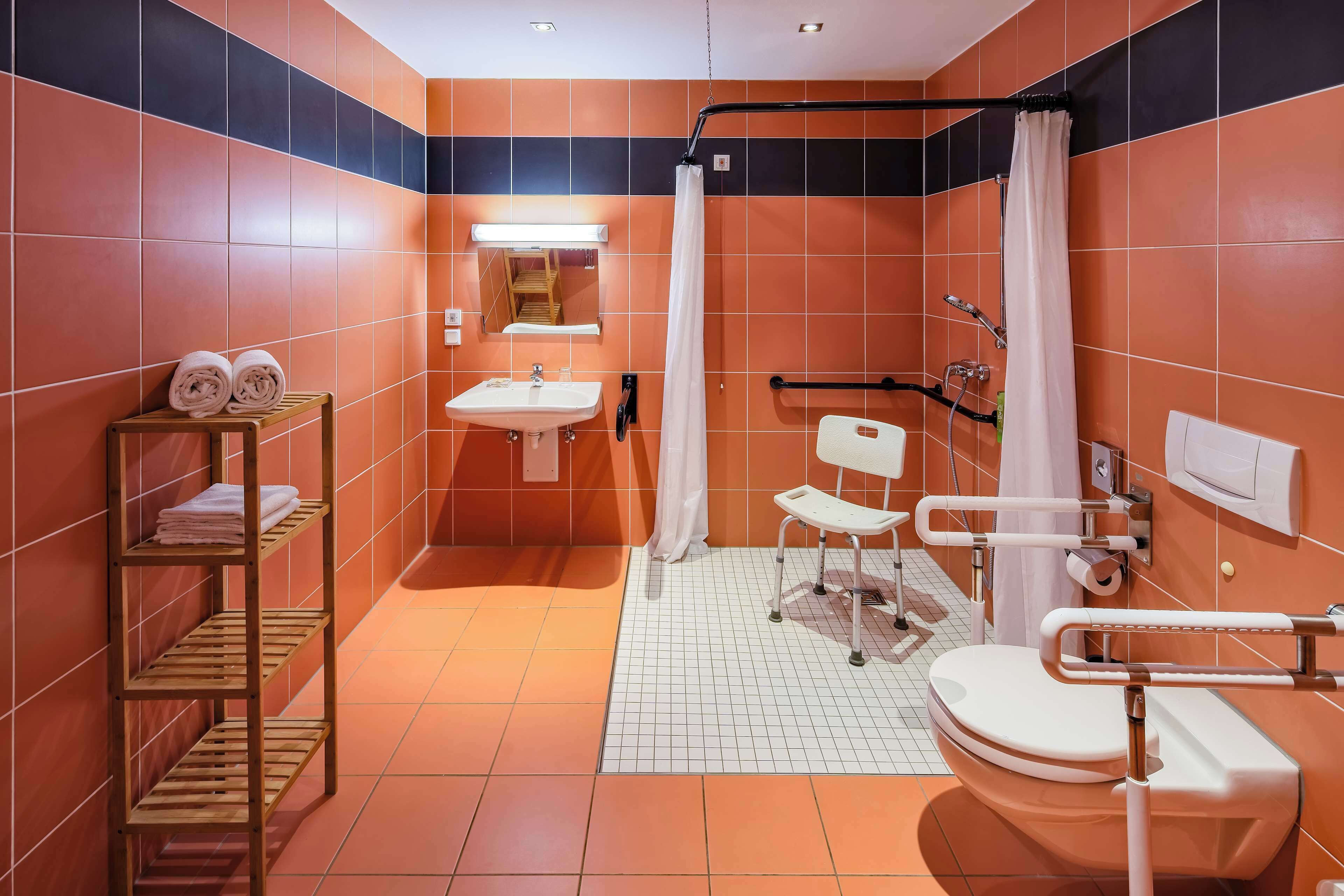 A bathroom with orange and black tiles, a white sink, a toilet, a shower with a white curtain, and a wooden shelf with towels.