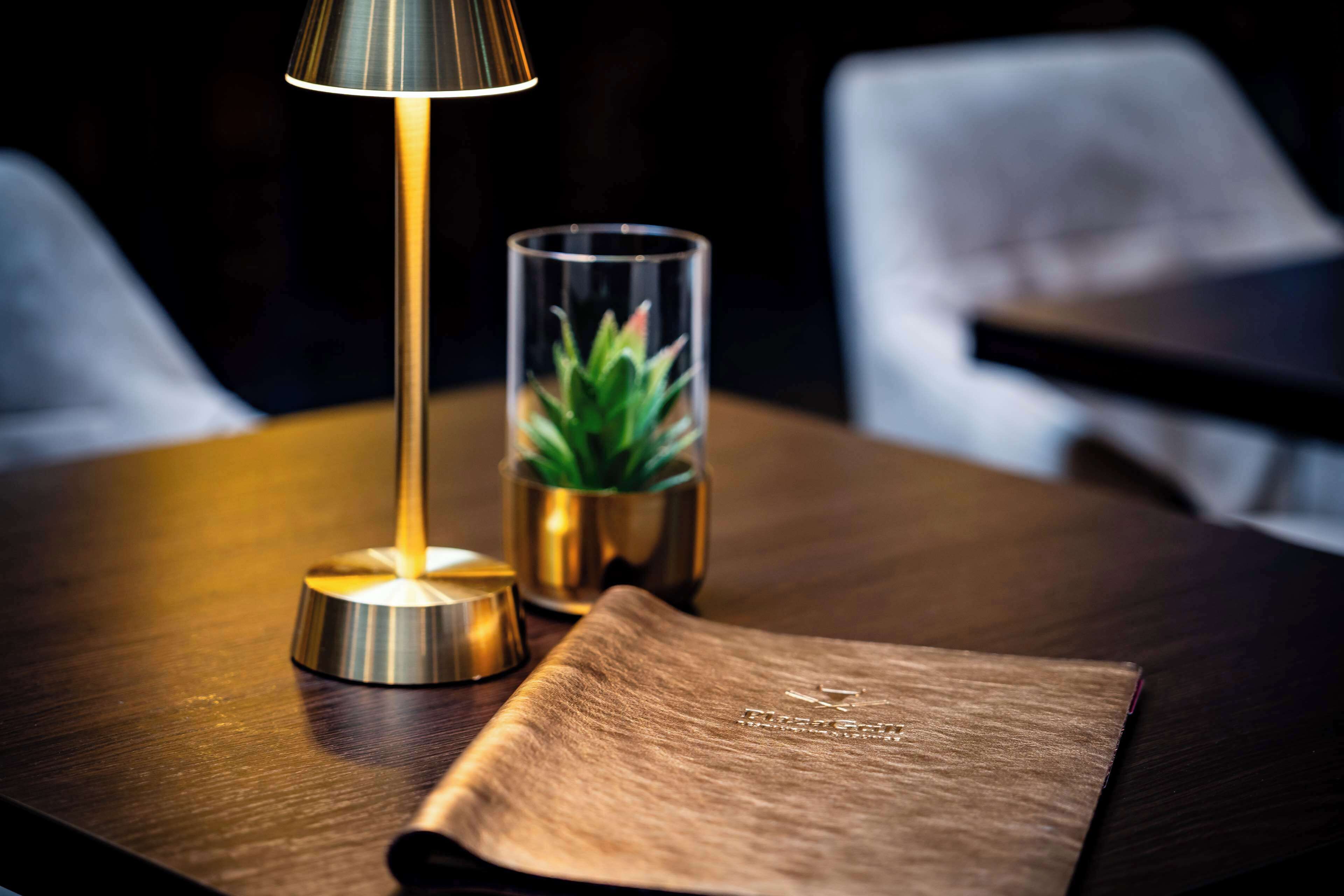 Uma mesa de madeira com uma luminária, um recipiente de vidro com uma planta e um caderno de couro.