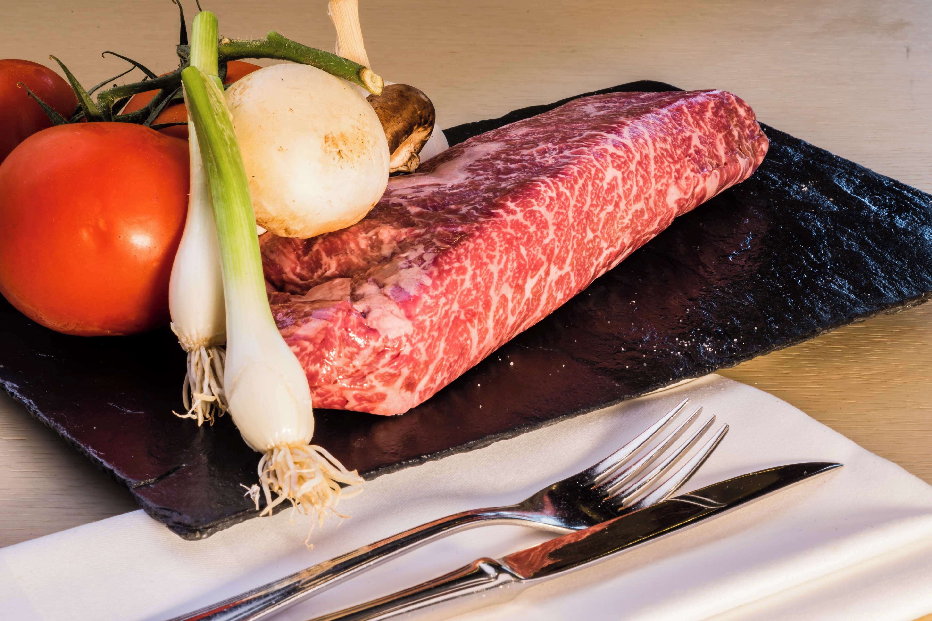 A raw steak, a tomato, a bulb of garlic, and a mushroom on a black slate plate.