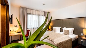 A modern hotel room with a large bed, a desk with a lamp, a chair, and a potted plant. - Park Plaza Trier (Trier)