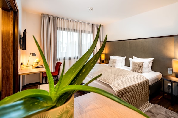 A modern hotel room with a large bed, a desk with a lamp, a chair, and a potted plant.