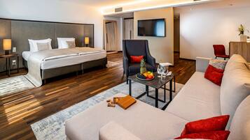 A modern hotel room with a bed, a sofa, a chair, a small table with fruit and a bottle, and a TV on the wall.
