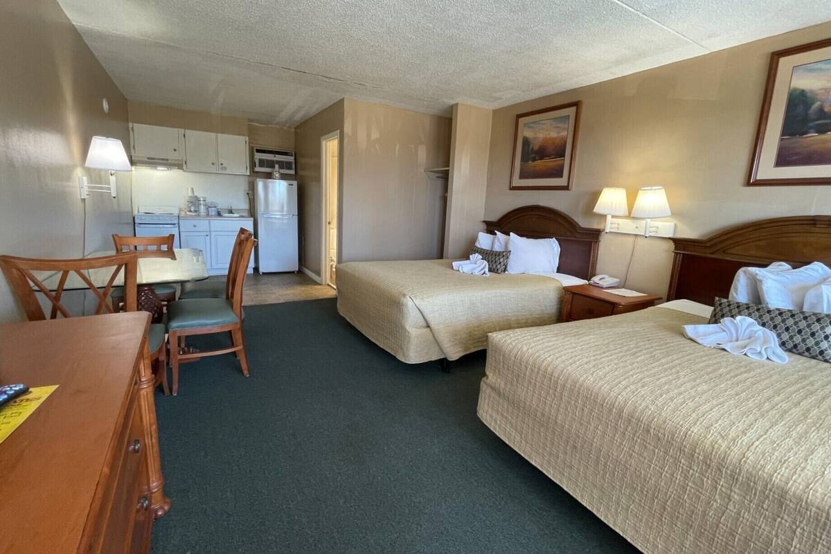 A hotel room with two beds, a desk, a chair, a small kitchenette, and a painting on the wall.