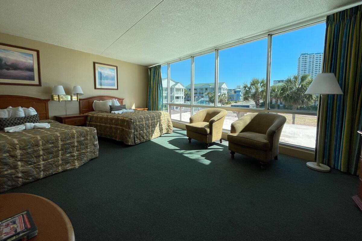 A hotel room with two beds, a sofa, and large windows offering a view of buildings and palm trees.