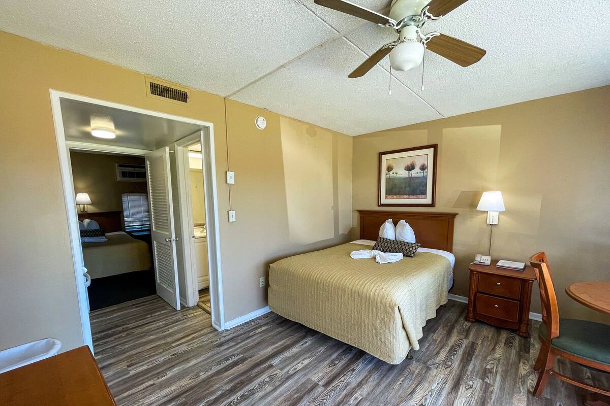 A hotel room with a bed, a desk, a chair, a ceiling fan, and a painting on the wall.