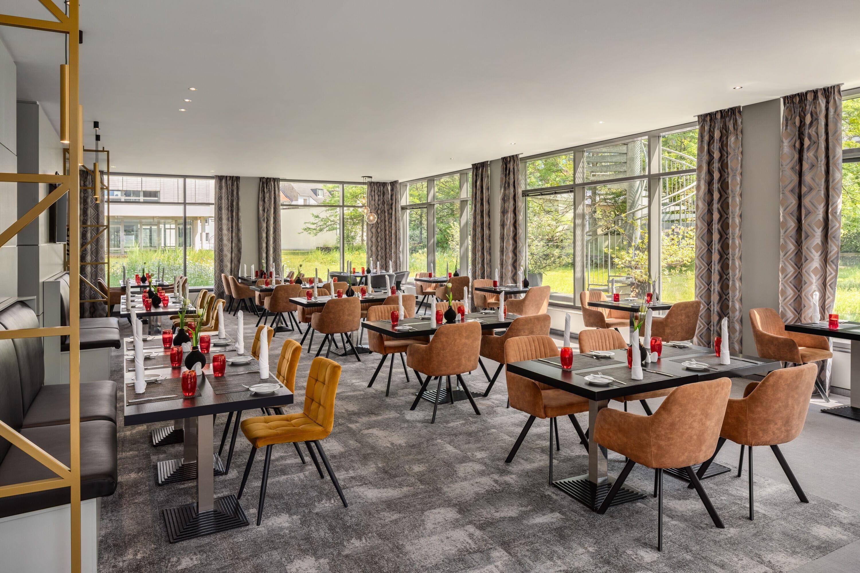 Un comedor amplio con múltiples mesas y sillas, ventanas grandes que ofrecen vistas a zonas verdes, y un diseño de interiores moderno.