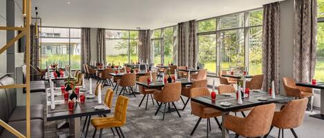 Un comedor amplio con múltiples mesas y sillas, ventanas grandes que ofrecen vistas a zonas verdes, y un diseño de interiores moderno.
