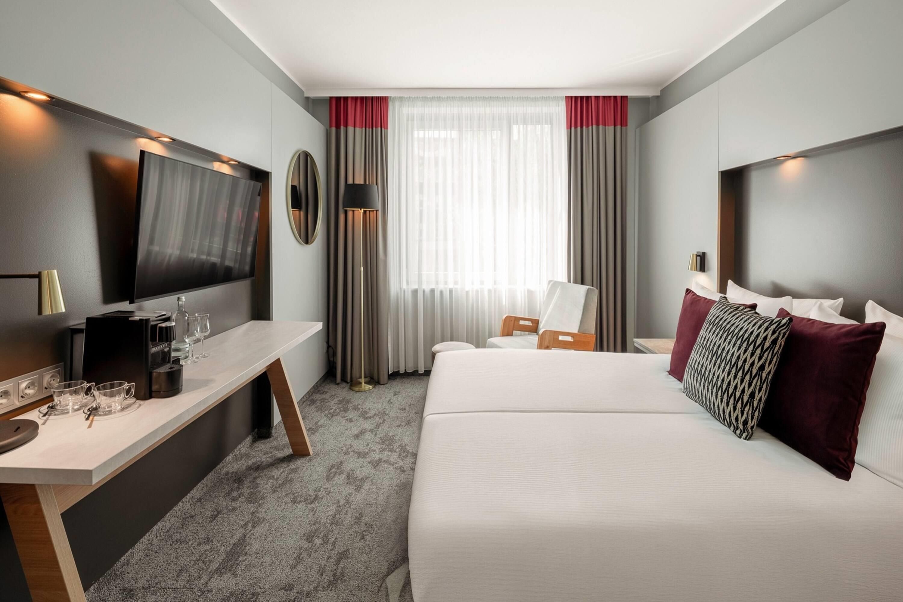 Habitación de hotel con cama, escritorio, televisión y ventana con cortinas.
