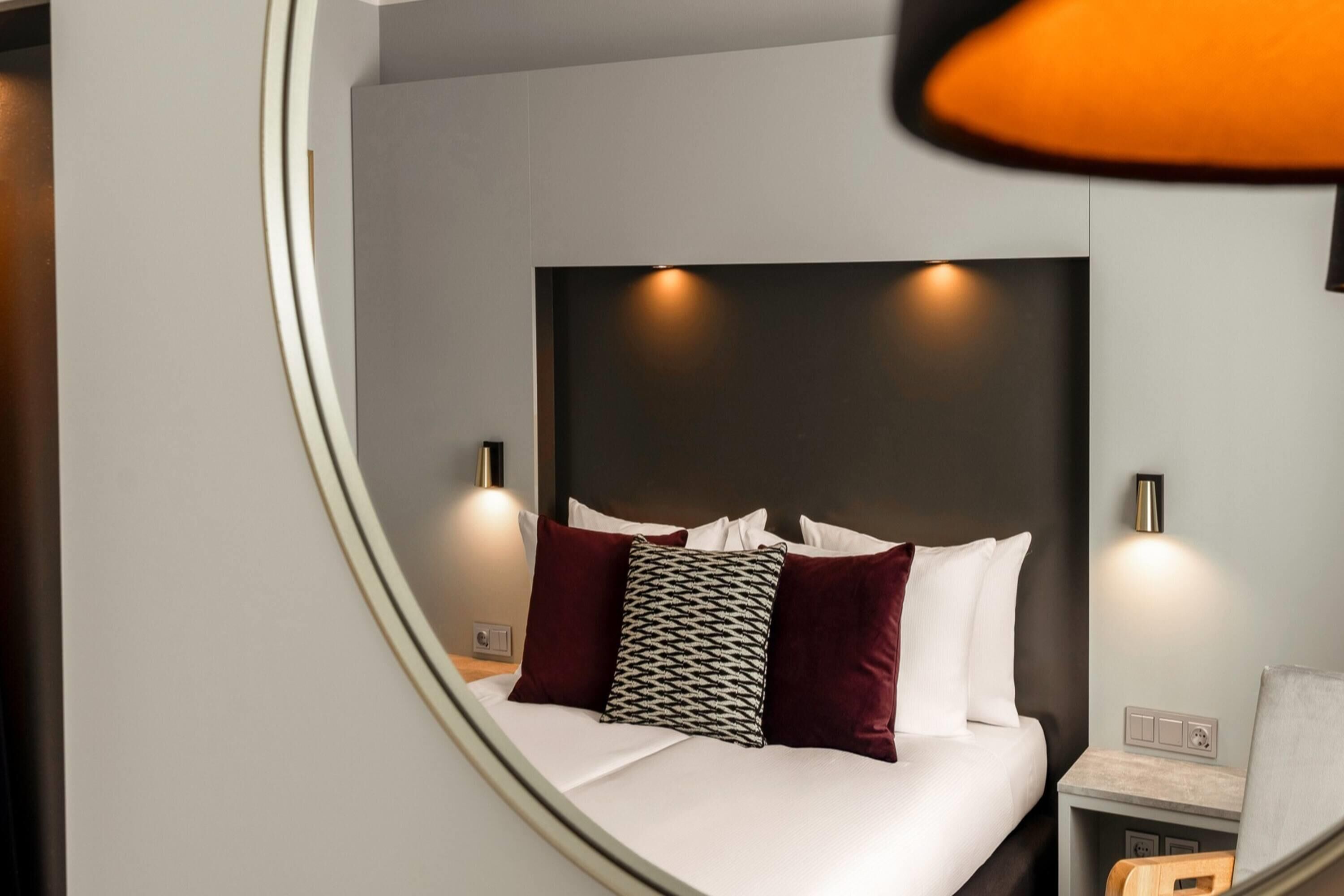 A modern hotel room with a curved bed, a large mirror, and a bedside table with a lamp.