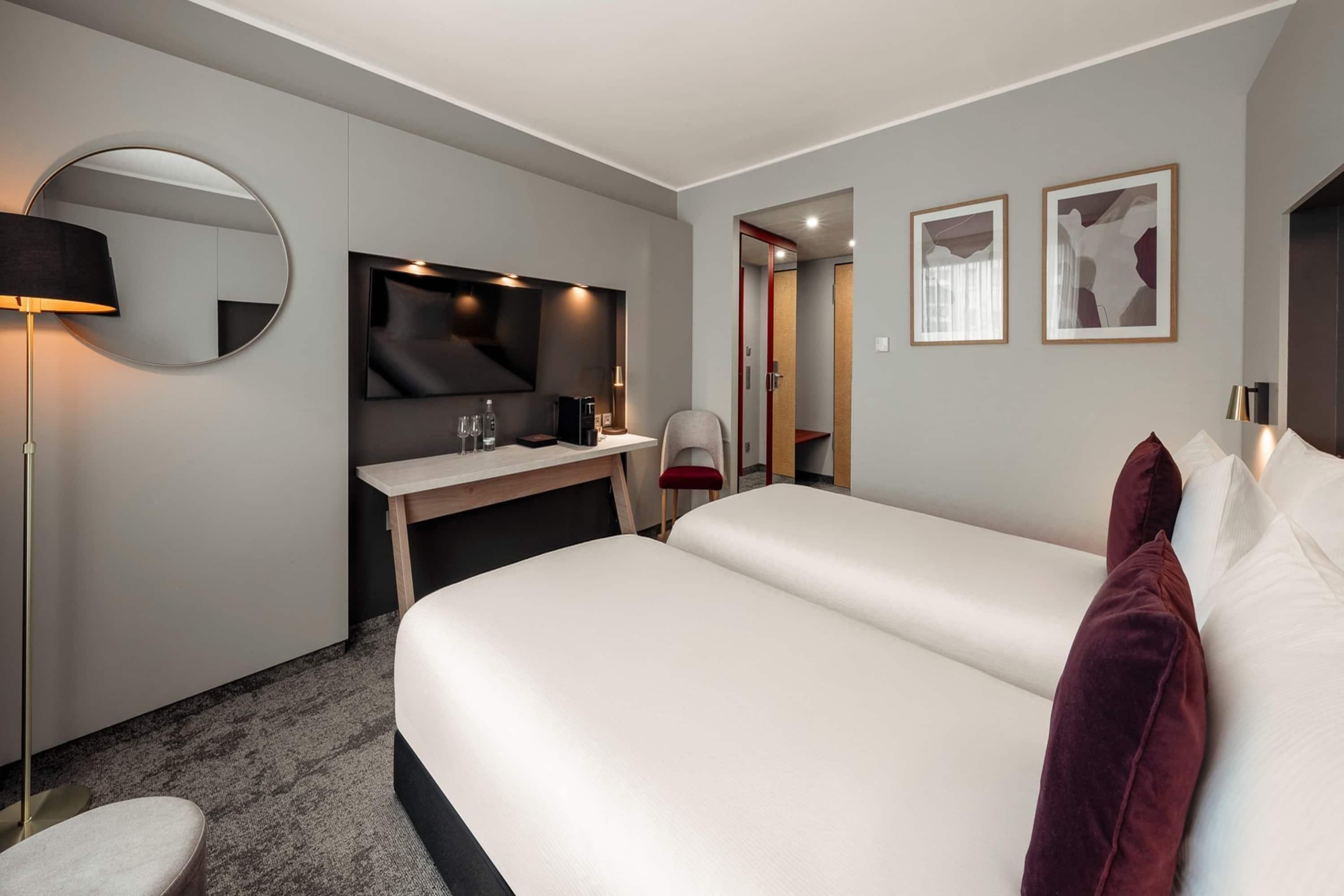 A hotel room with a large bed, a desk with a chair, a round mirror, a flat-screen TV, and two framed pictures on the wall.