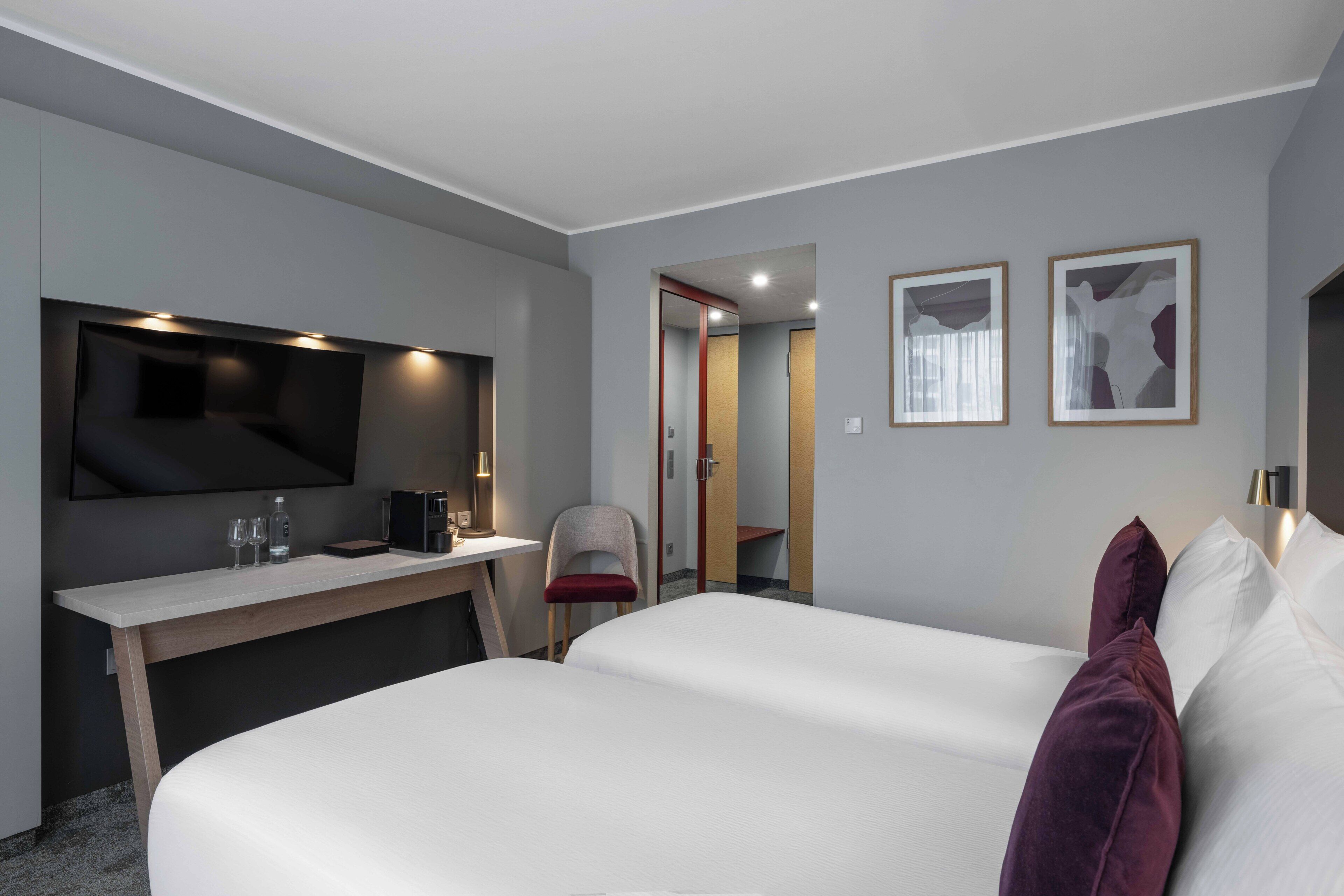 Ein Hotelzimmer mit einem Bett, einem Schreibtisch, einem Sessel, einem Fernseher und gerahmten Bildern an der Wand.