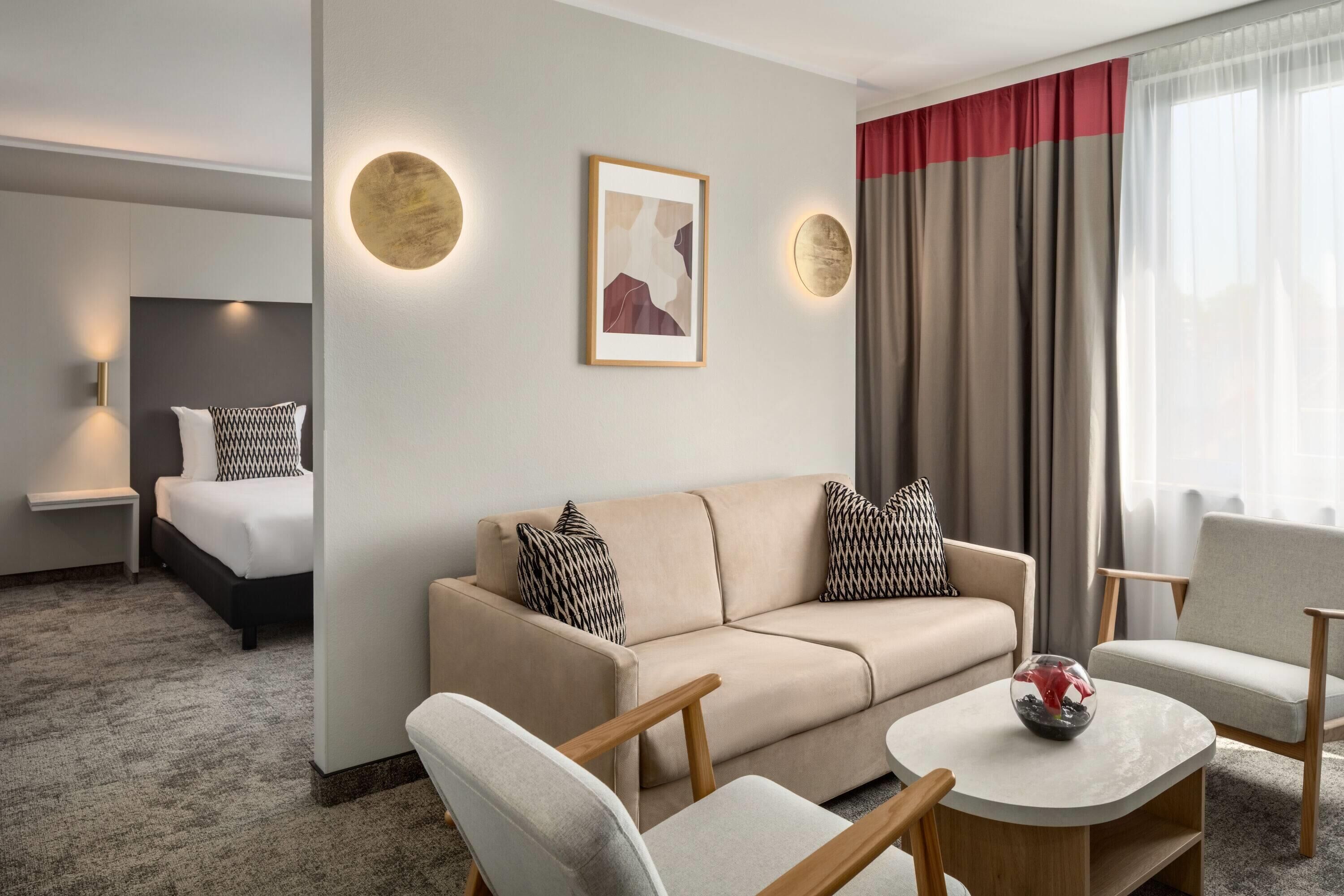 Ein Hotelzimmer mit einer Couch, Sesseln, einem kleinen Tisch, einem Bett und einem Bild an der Wand.