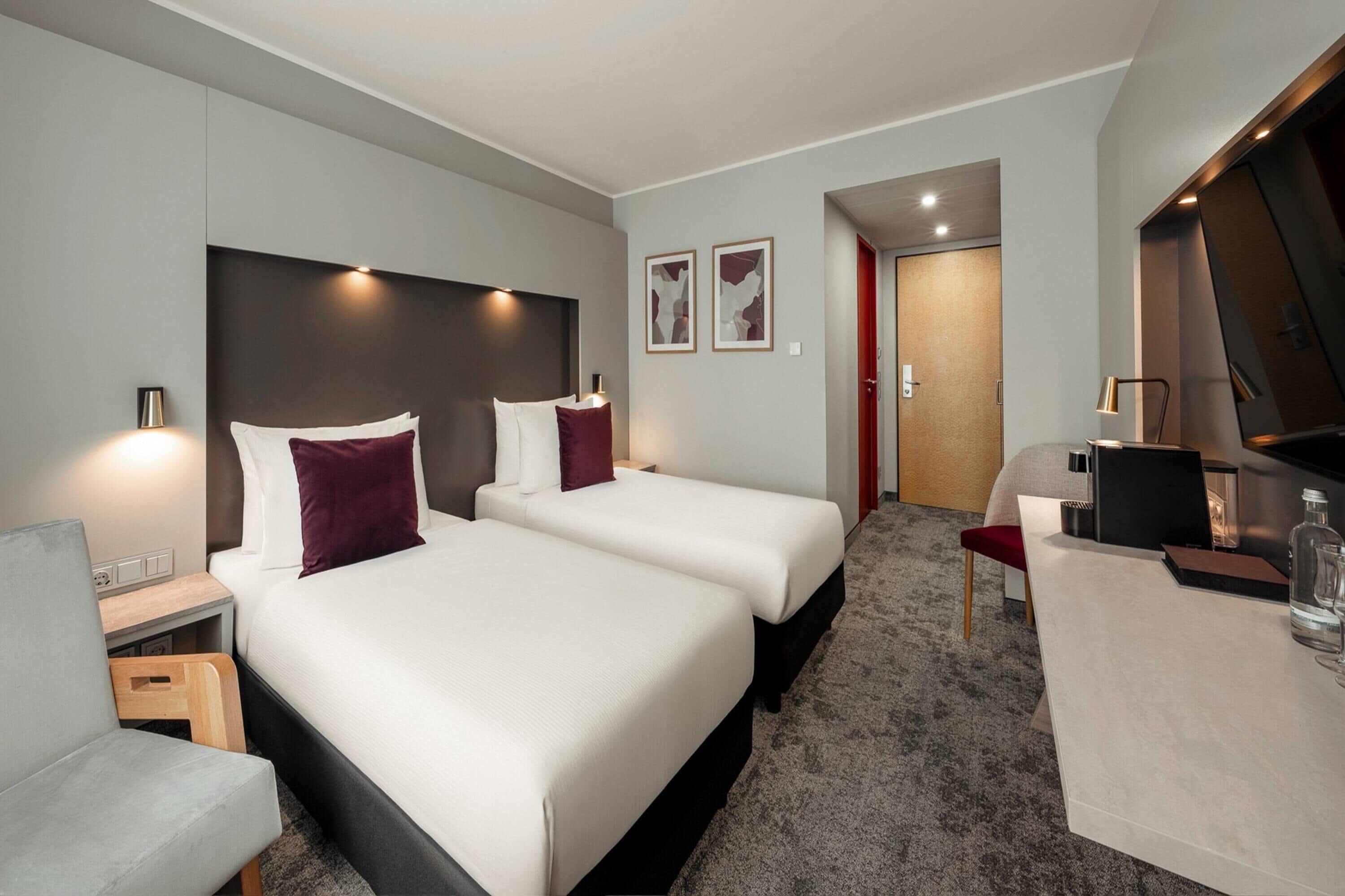 A hotel room with two beds, a desk, a chair, a TV, and a minibar.