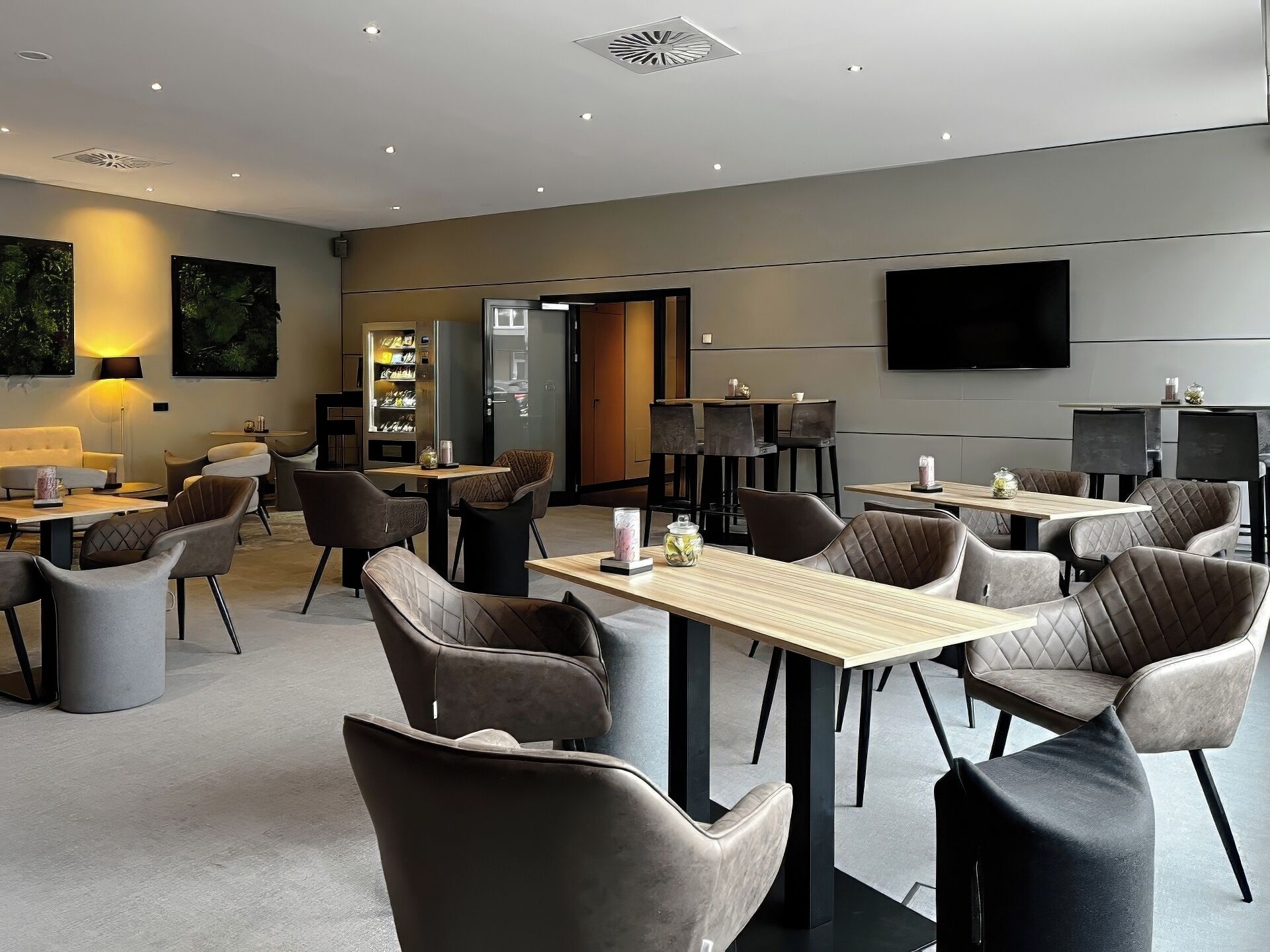 En moderne hotellobby med et centralt siddeområde, et køleskab med snacks og et stort fladskærms-tv på væggen.