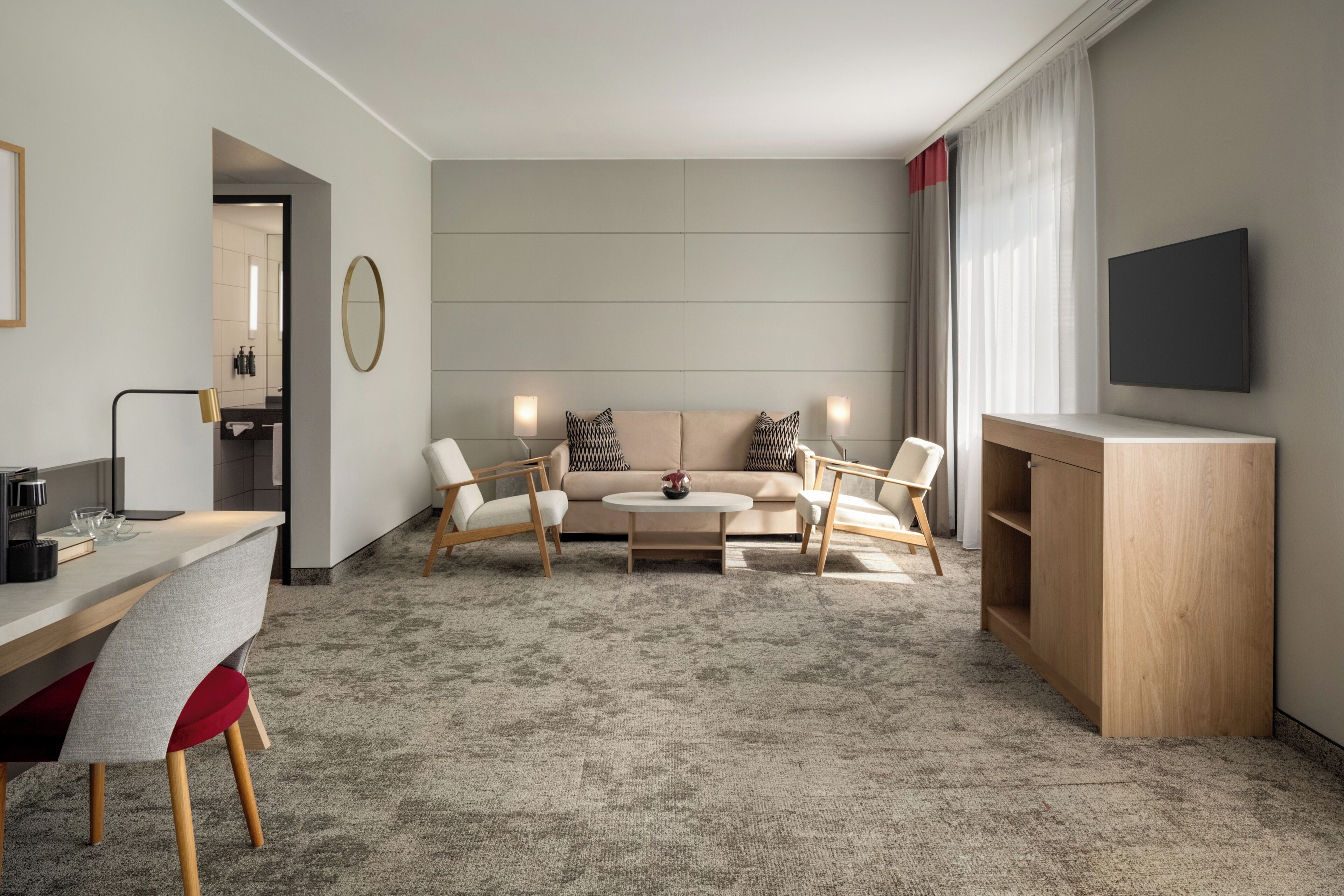 A modern hotel room with a sofa set, a coffee table, a desk with a chair, a television, and a large window with curtains.