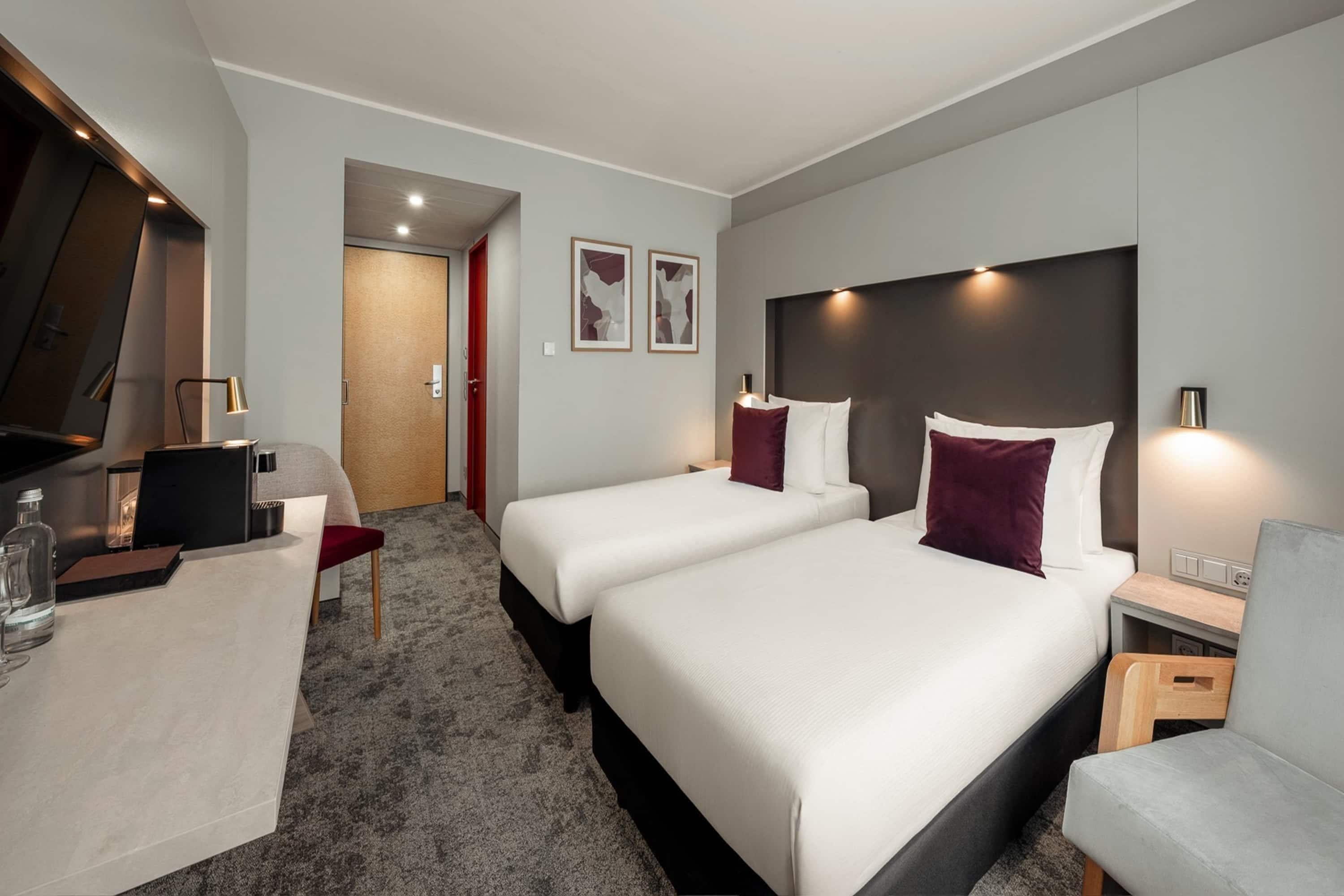 A hotel room with two beds, a desk, a chair, a TV, and a door leading to another room.