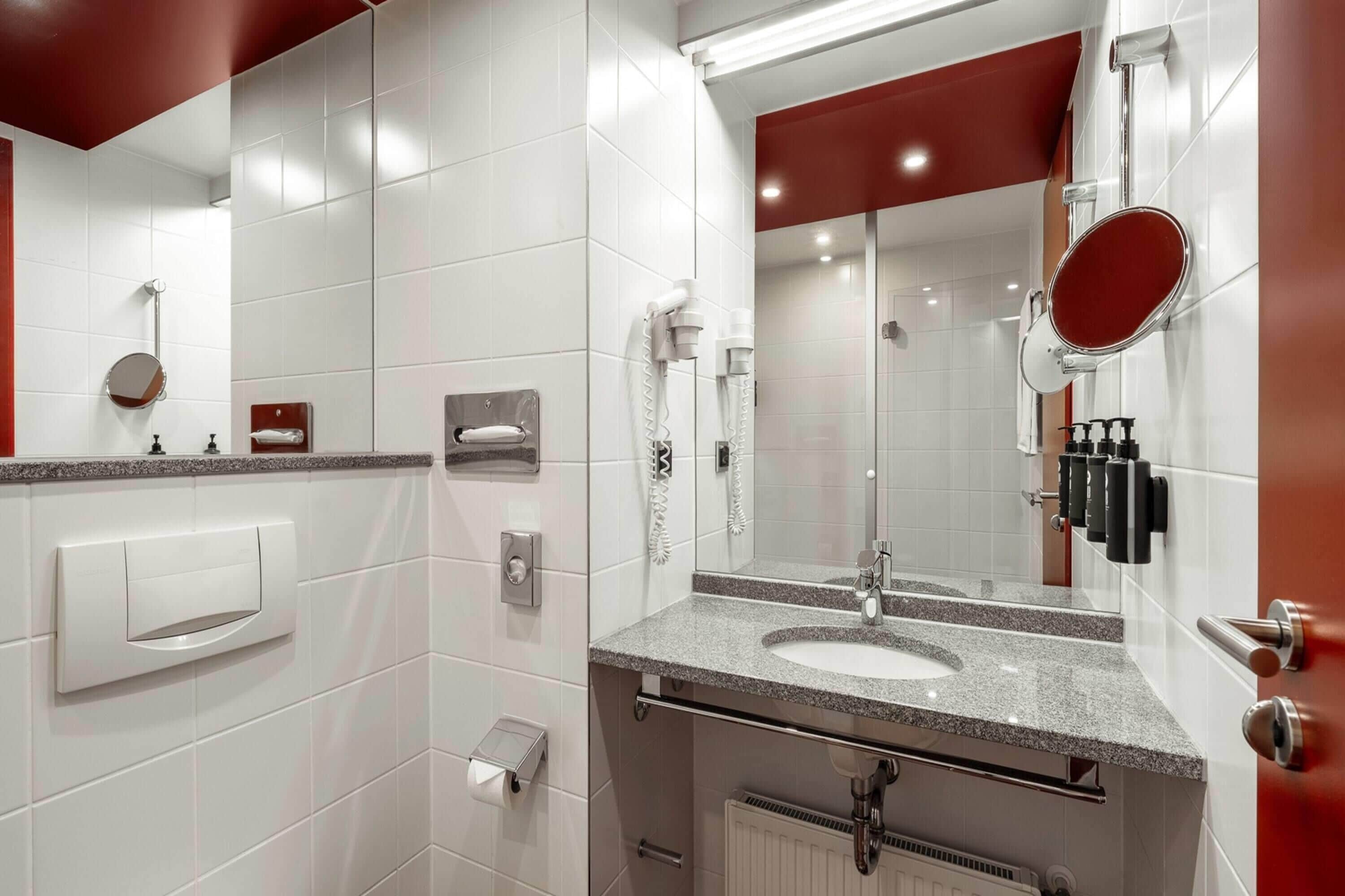 A modern bathroom with a white tiled wall, a granite countertop, a sink, a mirror, a towel dispenser, a toilet paper holder, and a shower area.