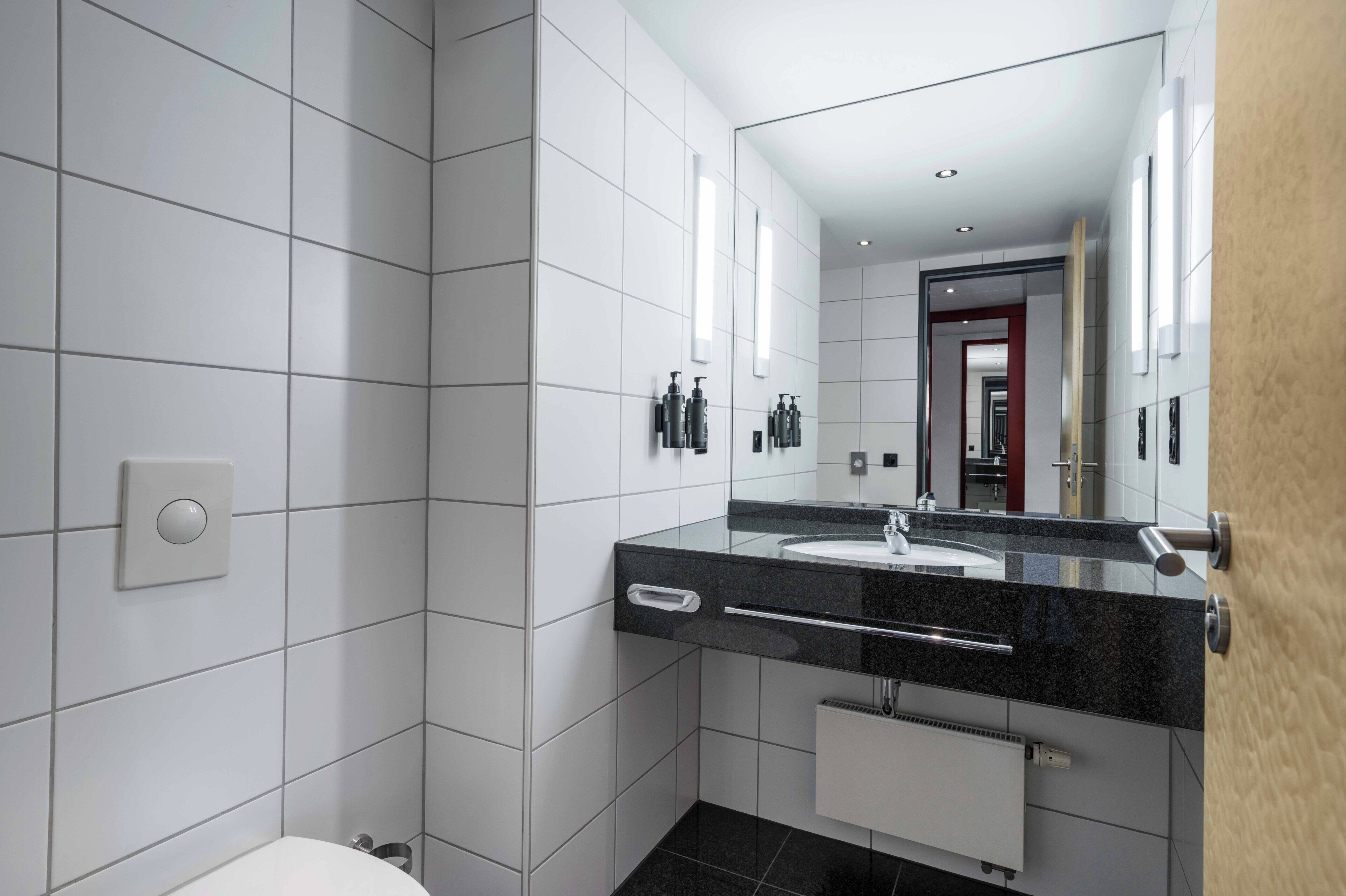 A modern bathroom with a large mirror, a black countertop, and a white tiled wall.