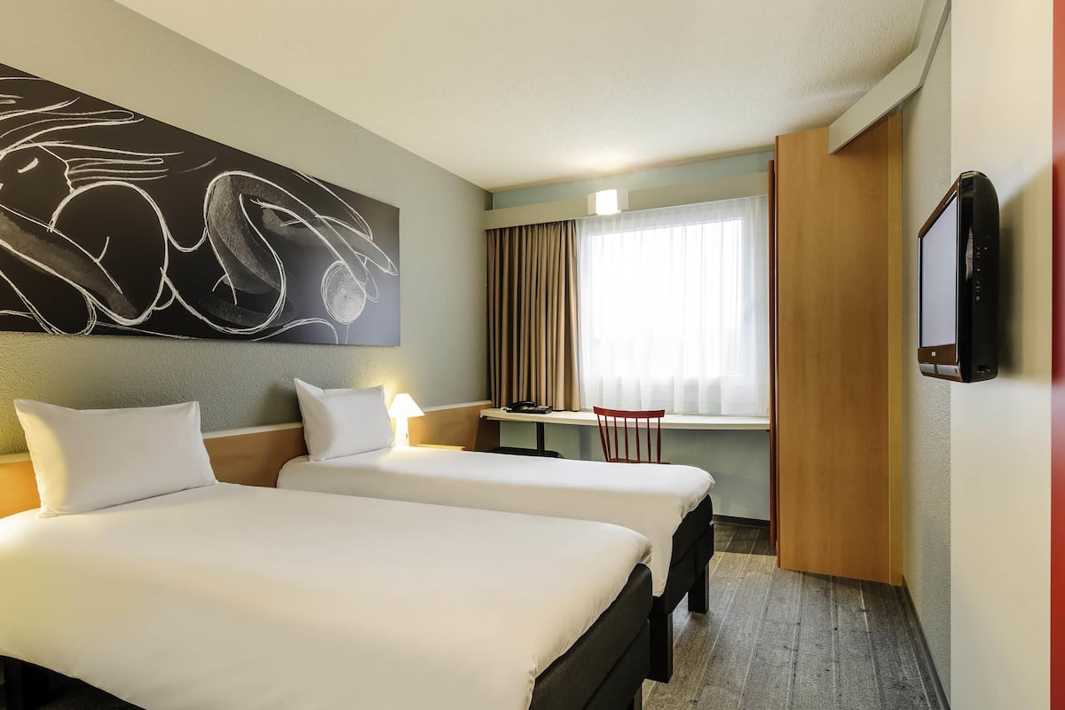 A hotel room with two beds, a desk, a chair, a TV, and a large abstract painting on the wall.
