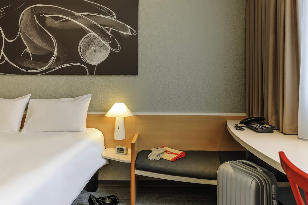 A hotel room with a bed, bedside table with a lamp, a telephone, a clock, a chair, a desk, and a suitcase.