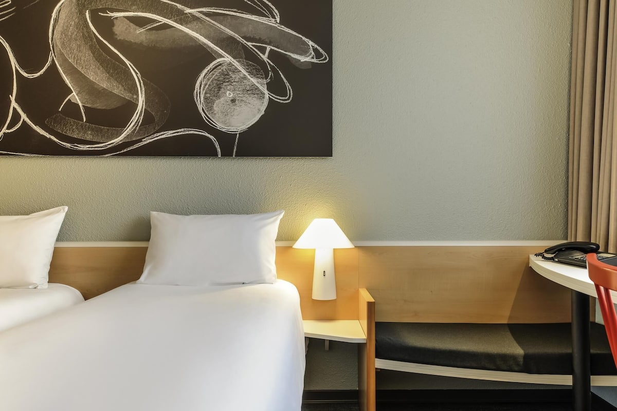 A hotel room with a bed, bedside table, lamp, chair, and a large abstract painting on the wall.