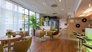Un interior moderno con suelo de madera, mesas redondas y sillas. Hay plantas en macetas y una zona de recepción con mostrador.