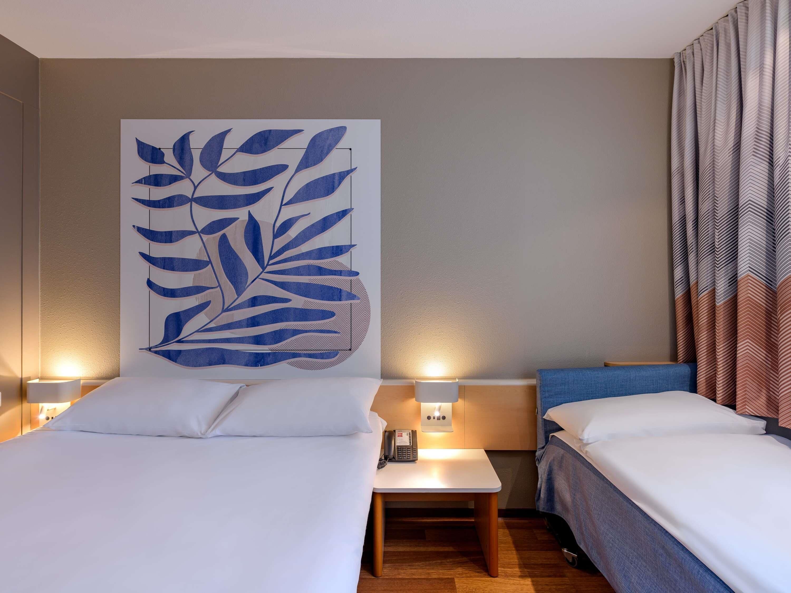 Habitación de hotel con dos camas, una mesita de noche con lámpara y una gran obra de arte abstracta en la pared.