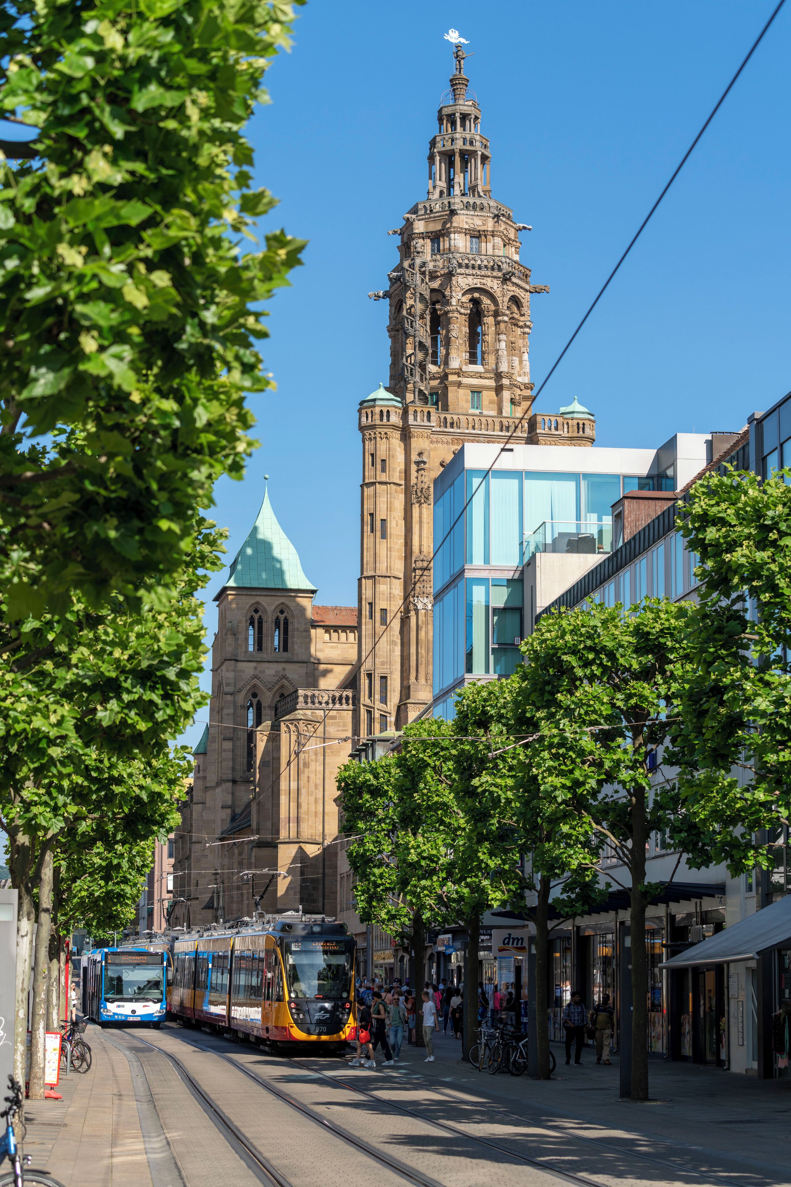Un tranvía pasando por un edificio histórico con una torre, rodeado de tiendas modernas y árboles.