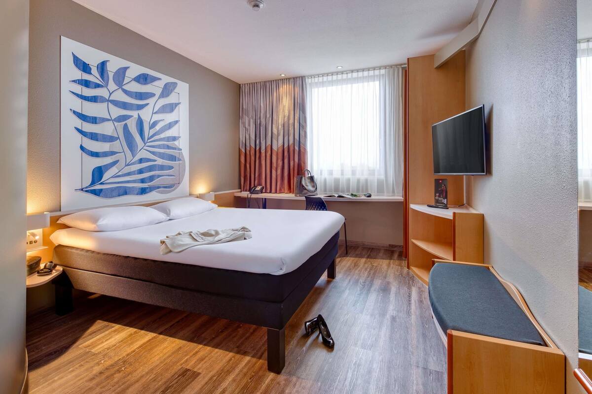 A hotel room with a bed, a desk with a chair, a TV, and a large window with curtains.