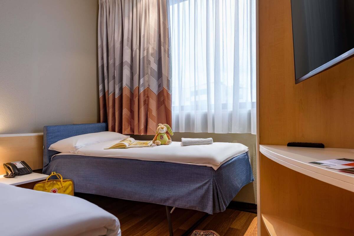A hotel room with a bed, a bedside table with a phone, a small table with a magazine, a window with curtains, and a TV on the wall.