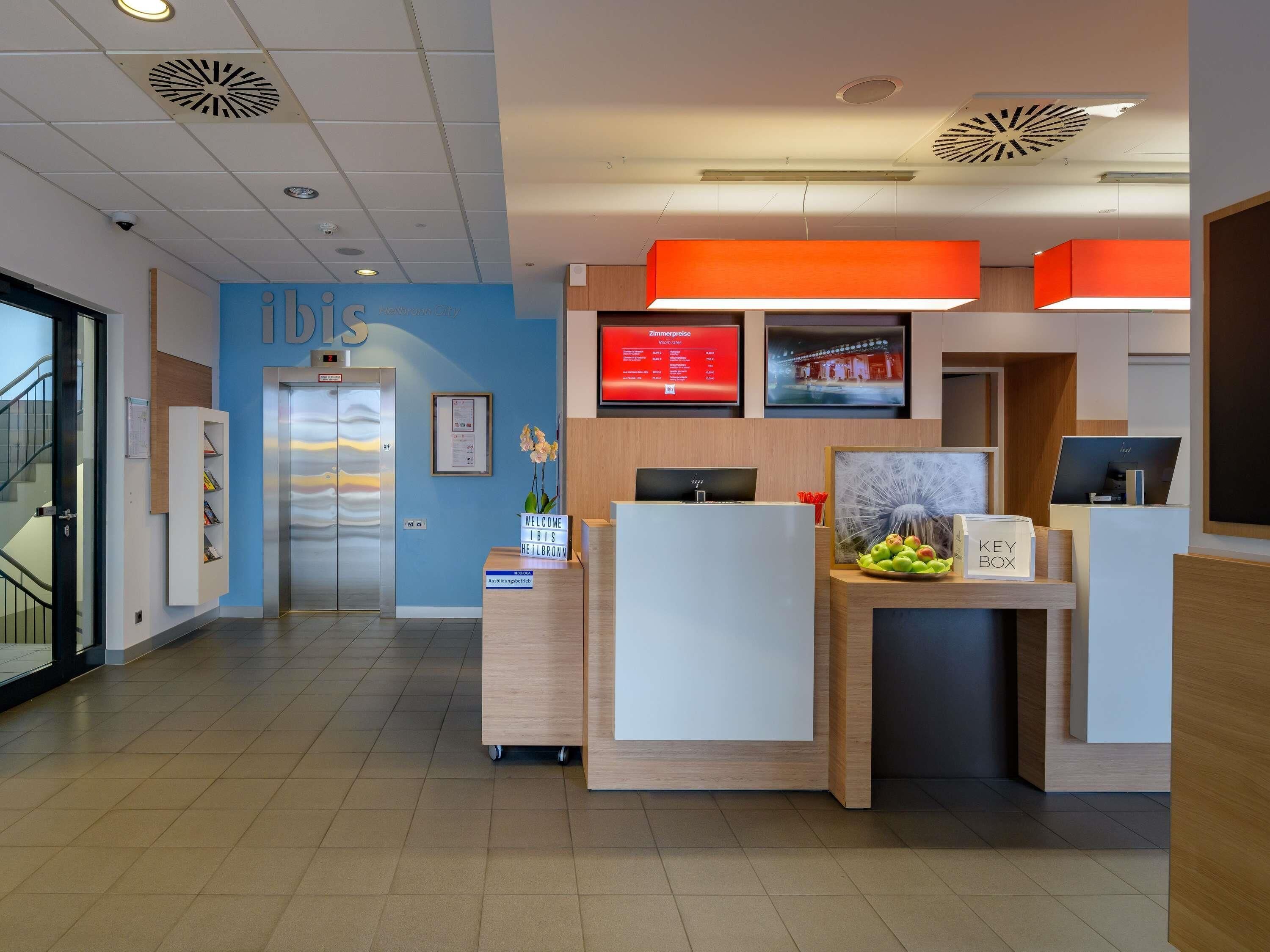 A modern hotel reception area with a blue wall, a reception desk, a digital display, and a staircase.