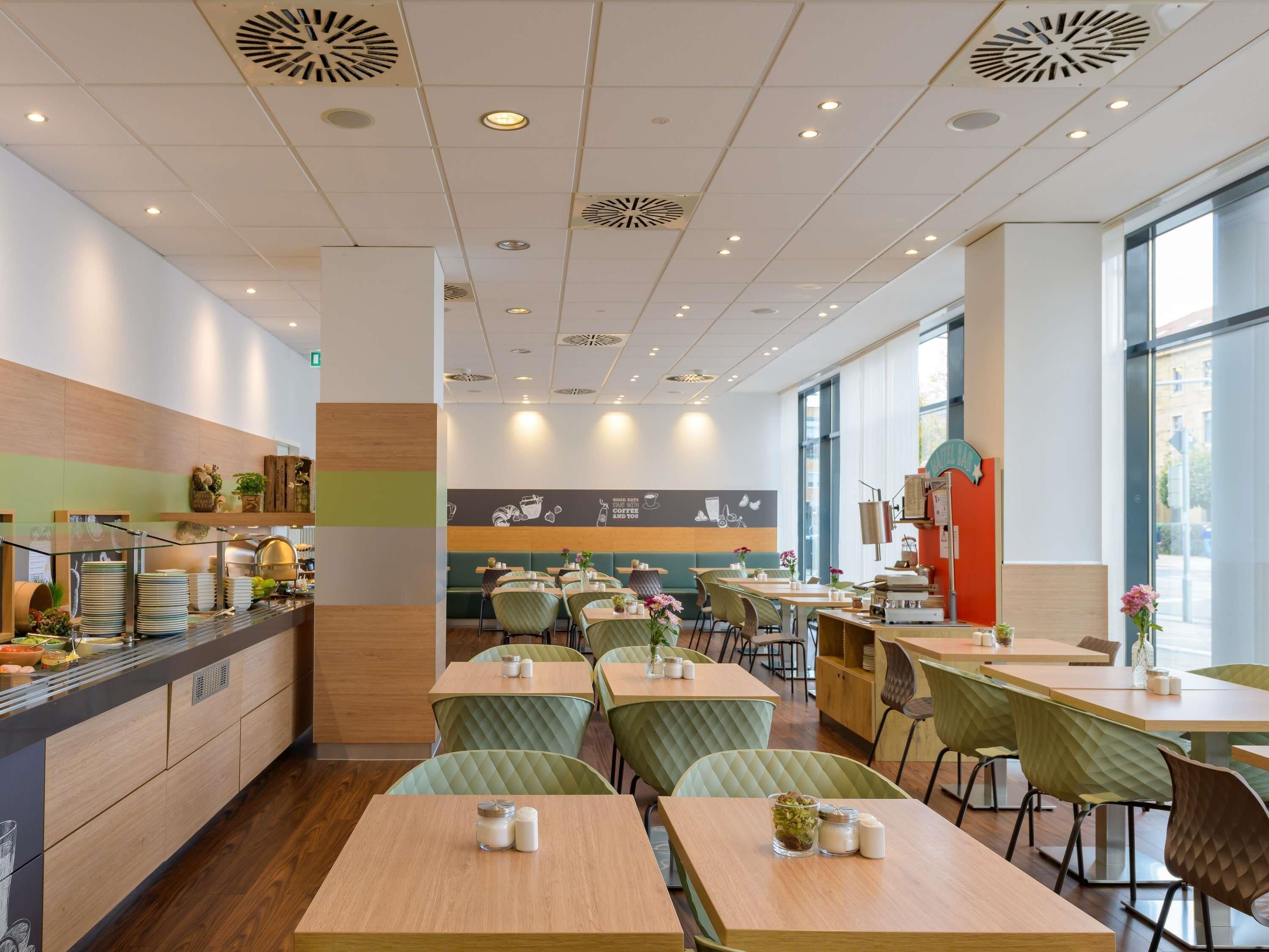 A modern cafe interior with wooden tables, green cushioned chairs, and a kitchen area with a counter.