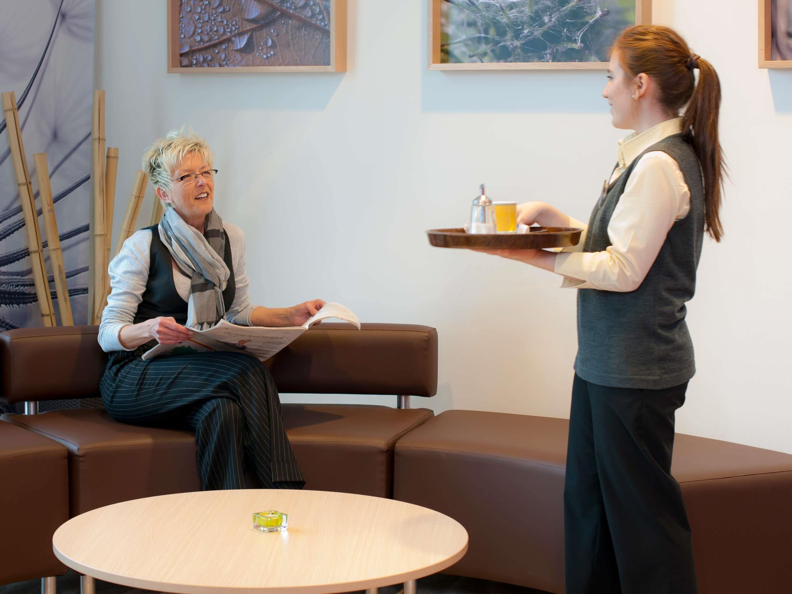 Une personne assise sur un canapé marron, en train de lire un livre, une autre personne debout, tenant un plateau de boissons, et une table ronde sur laquelle se trouve un petit objet en verre.