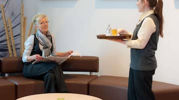 Une personne assise sur un canapé marron, en train de lire un livre, une autre personne debout, tenant un plateau de boissons, et une table ronde sur laquelle se trouve un petit objet en verre.