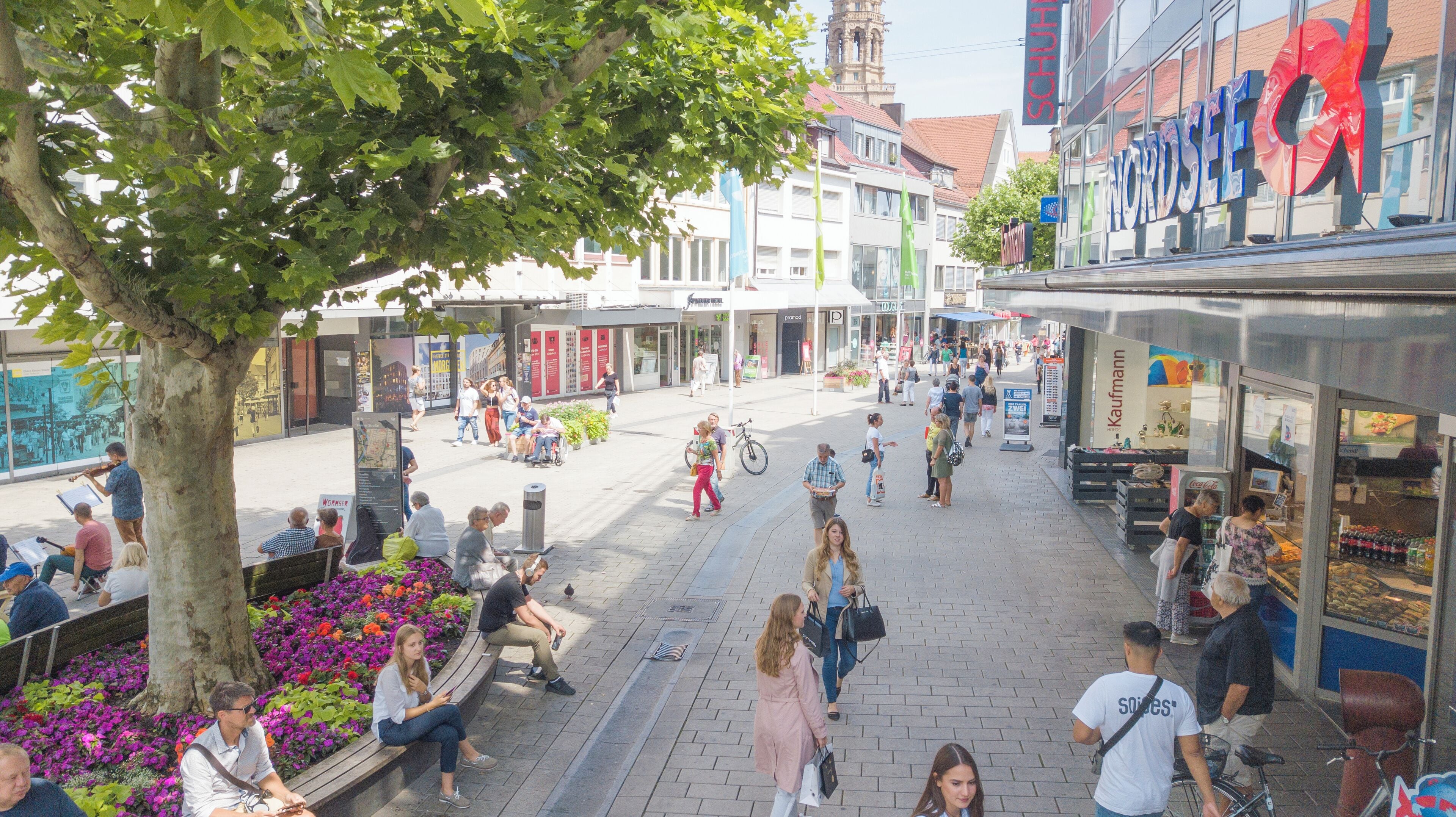 A bustling pedestrian street with shops and cafes, a large tree providing shade, and people walking and sitting.