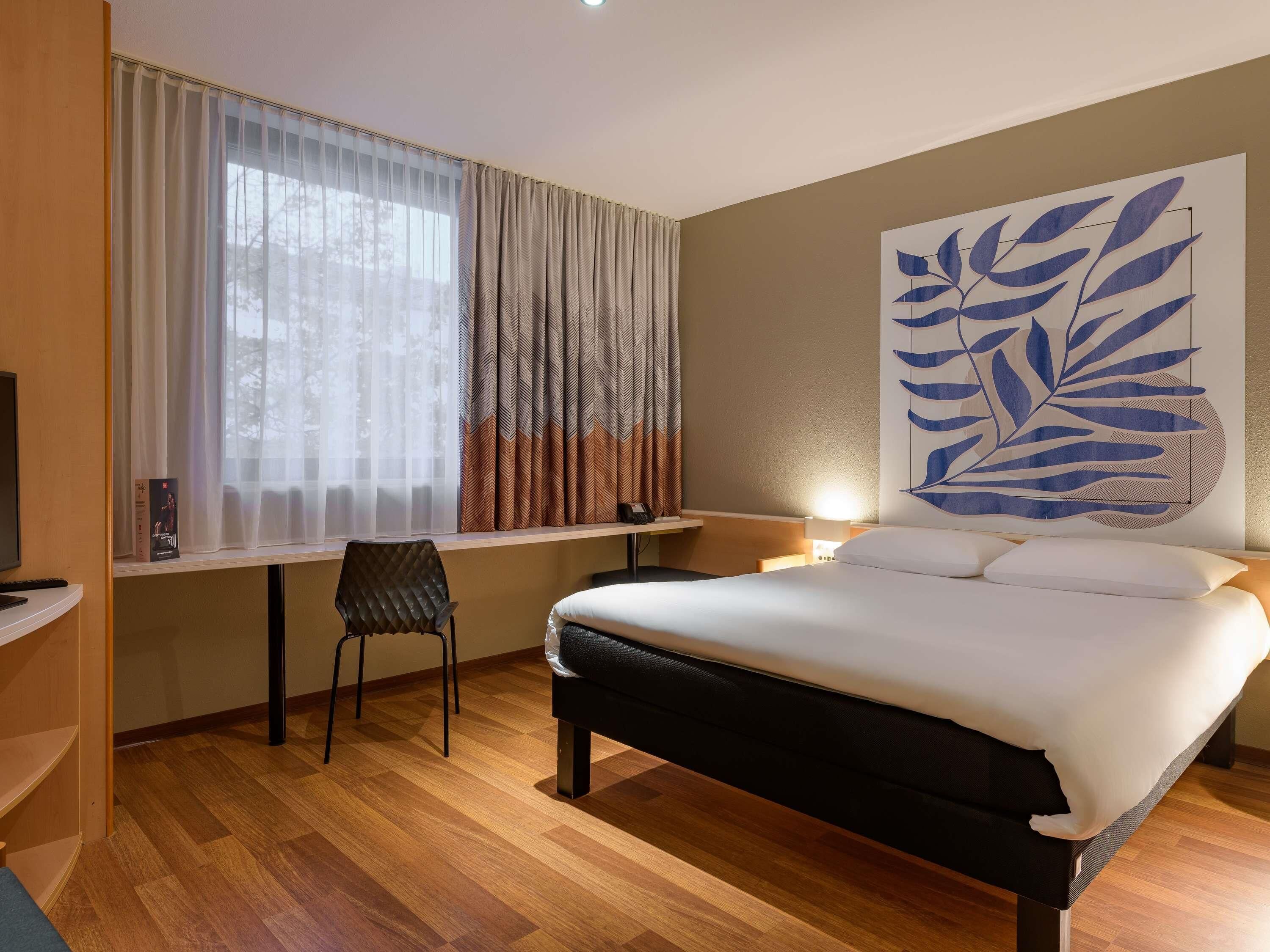 A hotel room with a bed, a desk, a chair, and a large leaf-patterned wall art.