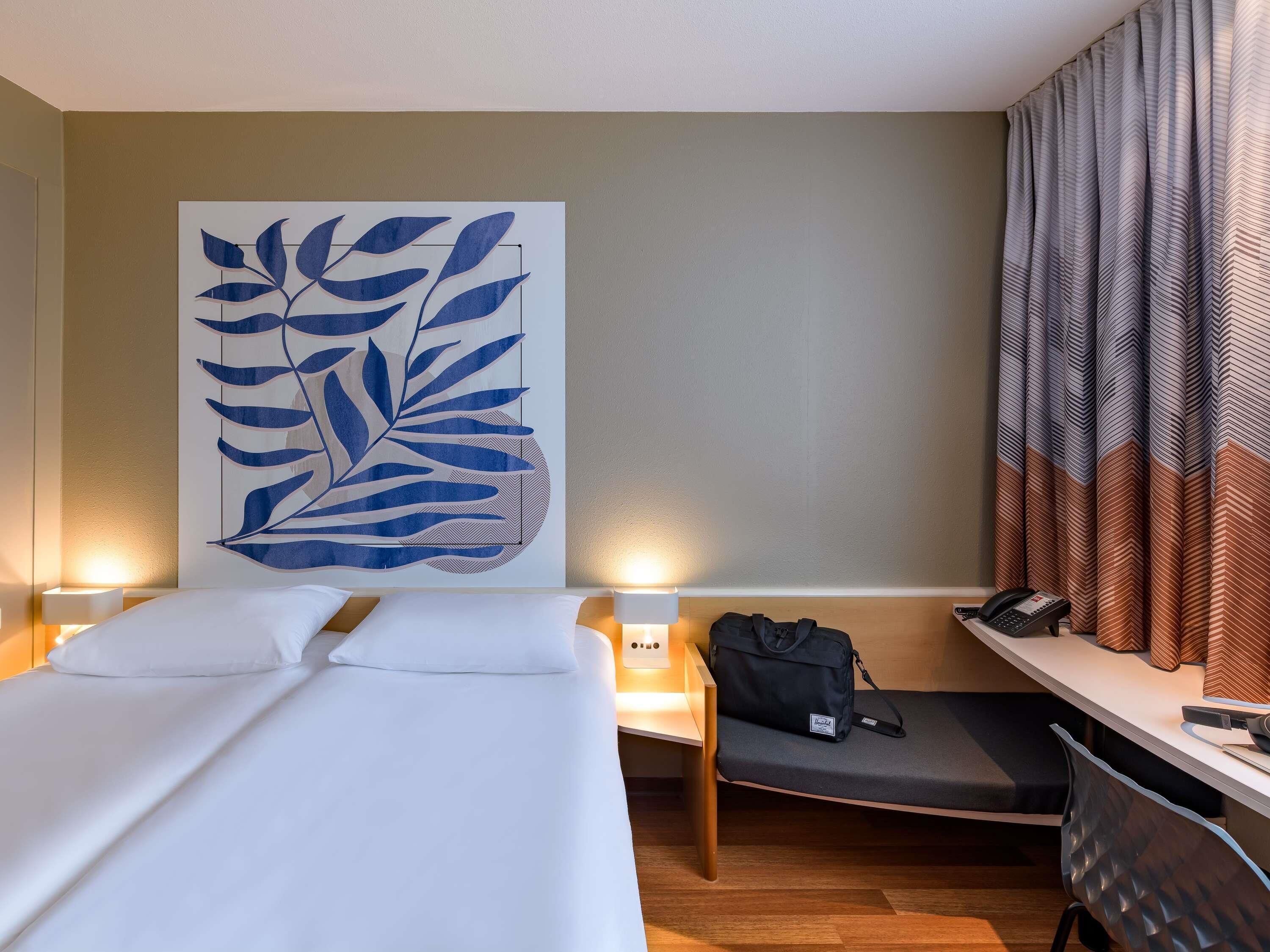 Habitación de hotel con cama, mesita de noche, lámpara, escritorio con silla y un cuadro grande con motivos de hojas en la pared.