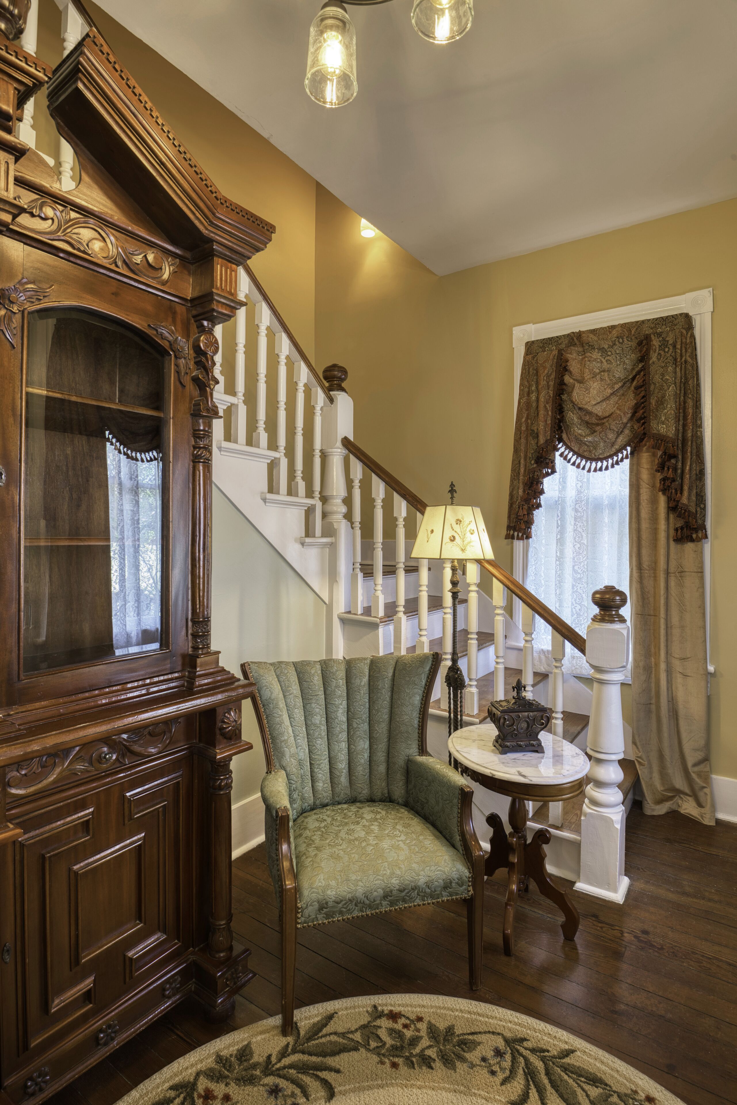 Una escalera con balaustres blancos y una barandilla de madera, una silla tapizada de verde, un mueble de madera y una mesa con una lámpara.