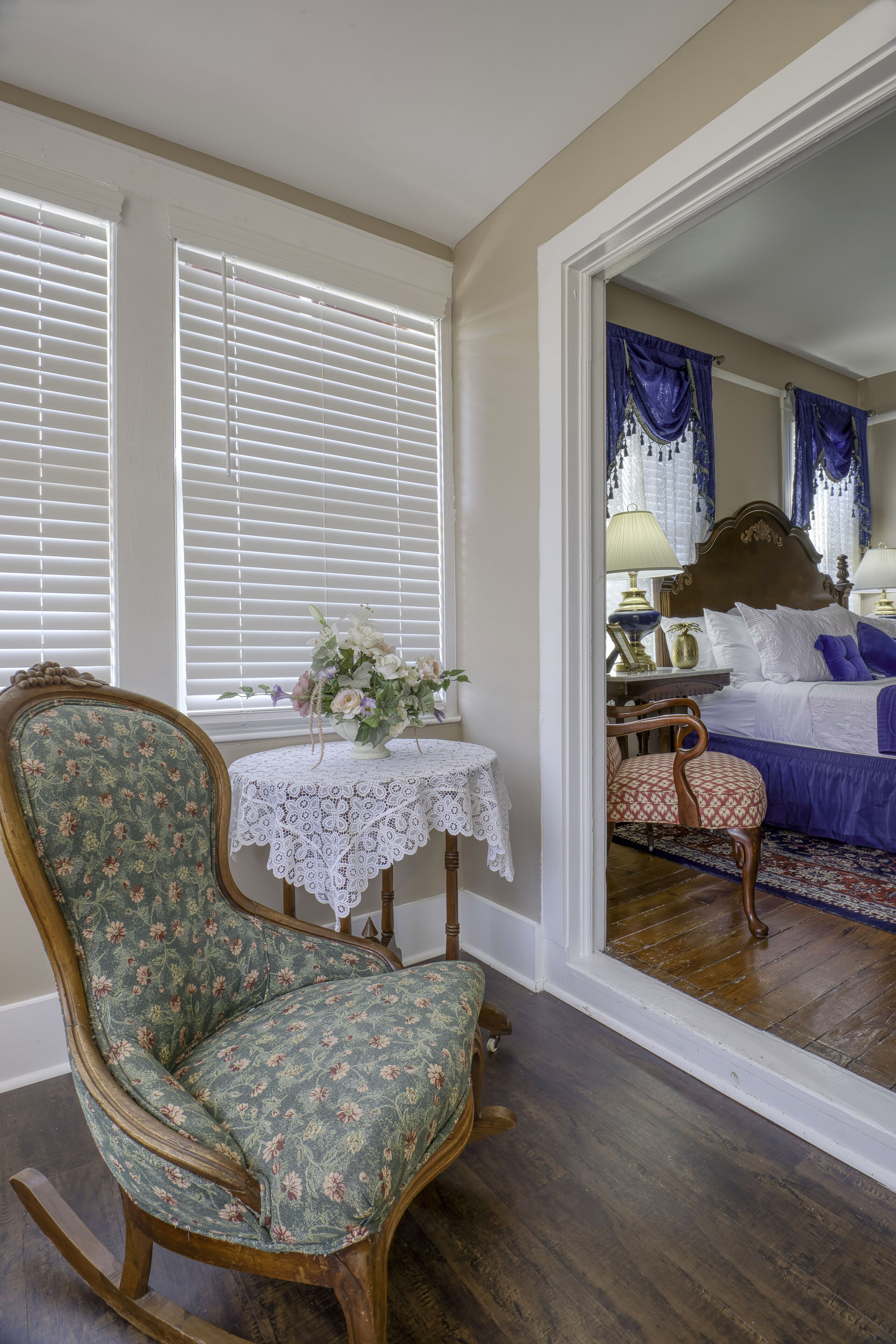 A room with a wooden rocking chair, a lace table, and a bed with blue curtains.