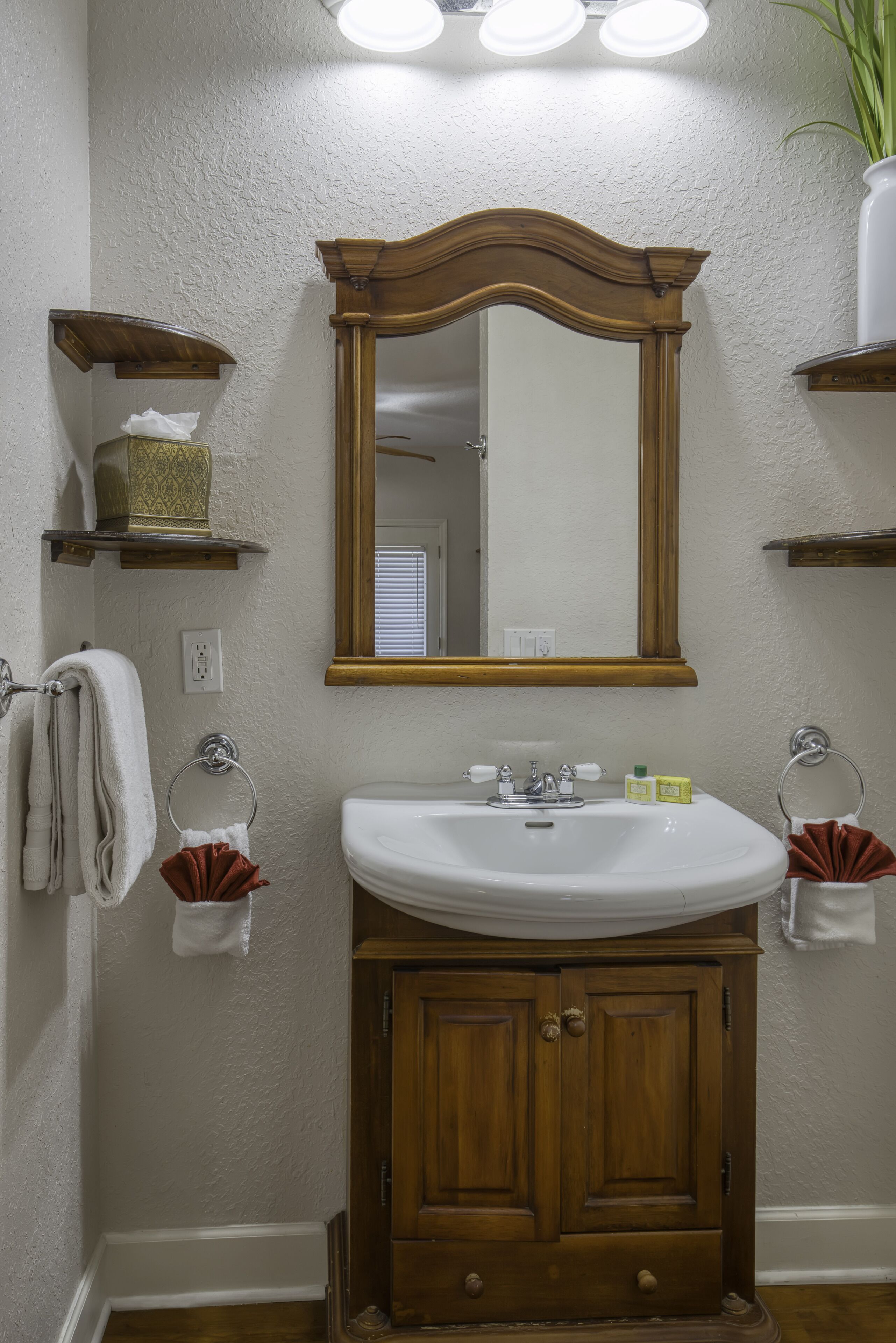 A bathroom with a wooden vanity, a white sink, and a mirror above it. There are two towel racks with towels and a small shelf with toiletries.