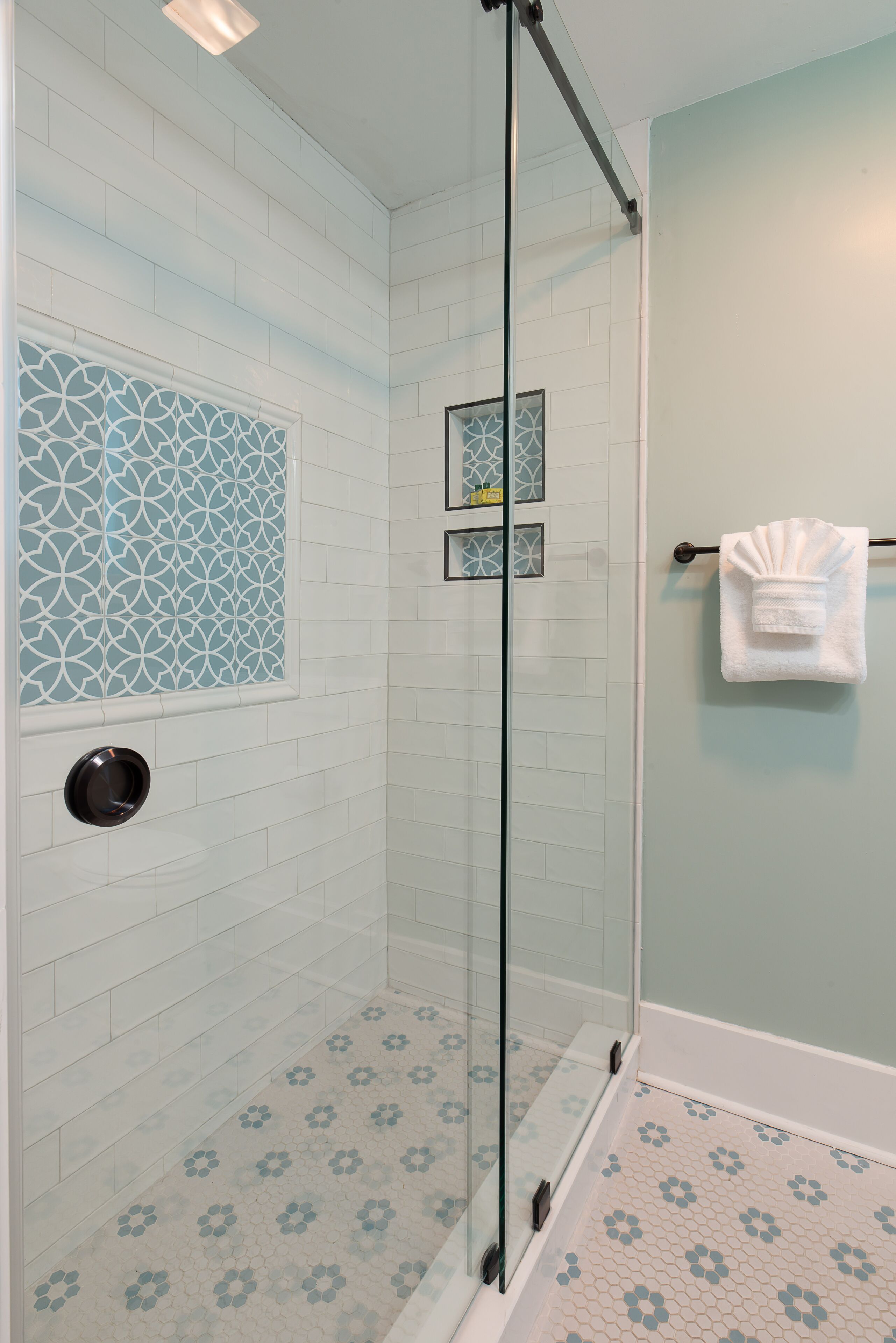 Una ducha con puertas de vidrio, paredes de azulejos blancos y suelo con diseño.