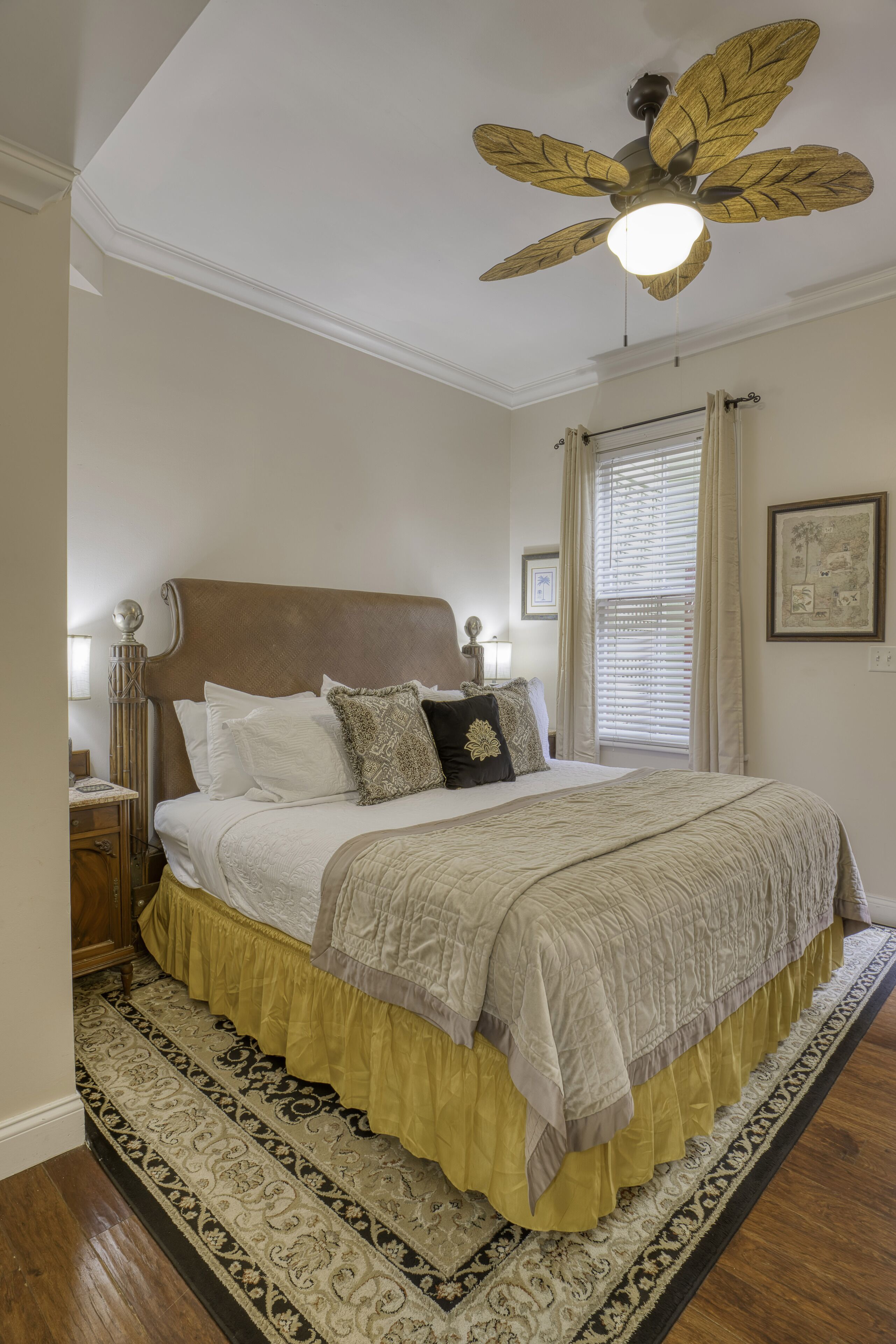 A bedroom with a large bed, a ceiling fan, a nightstand, a window with blinds, and a framed picture on the wall.