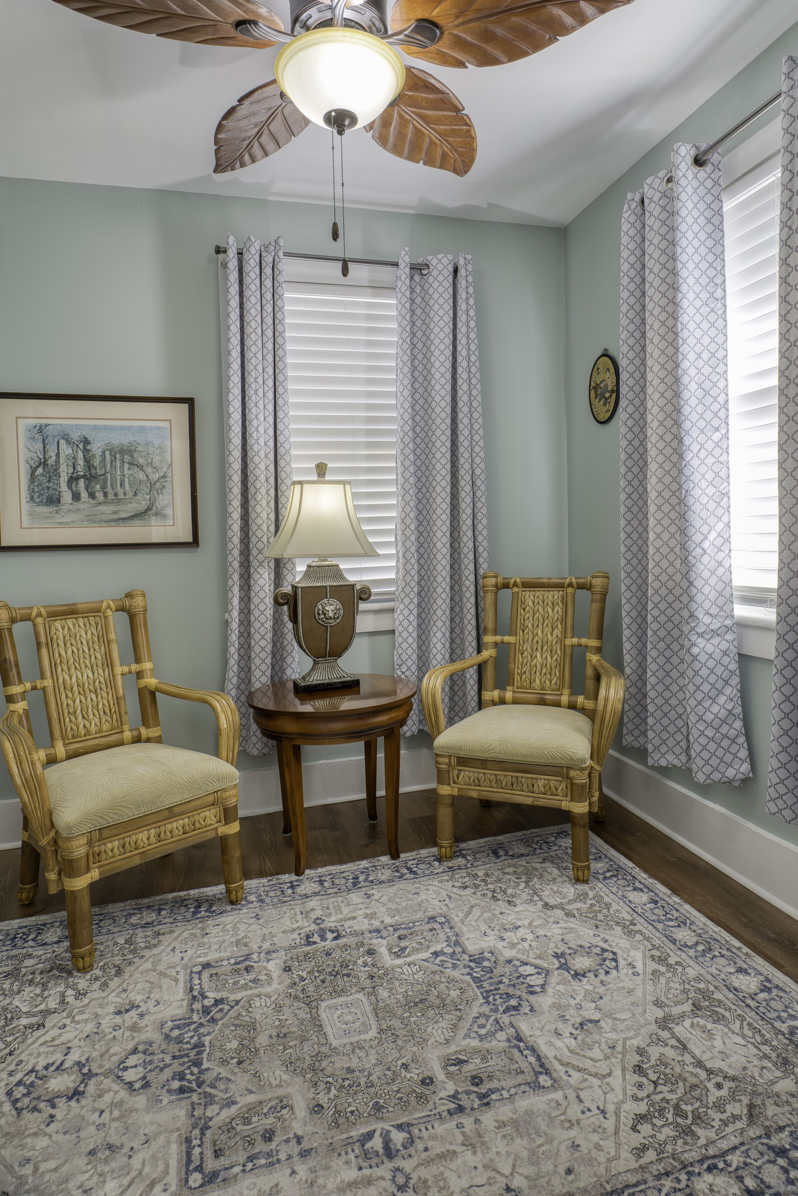 A room with a ceiling fan, two wicker chairs, a patterned rug, and a table with a lamp.